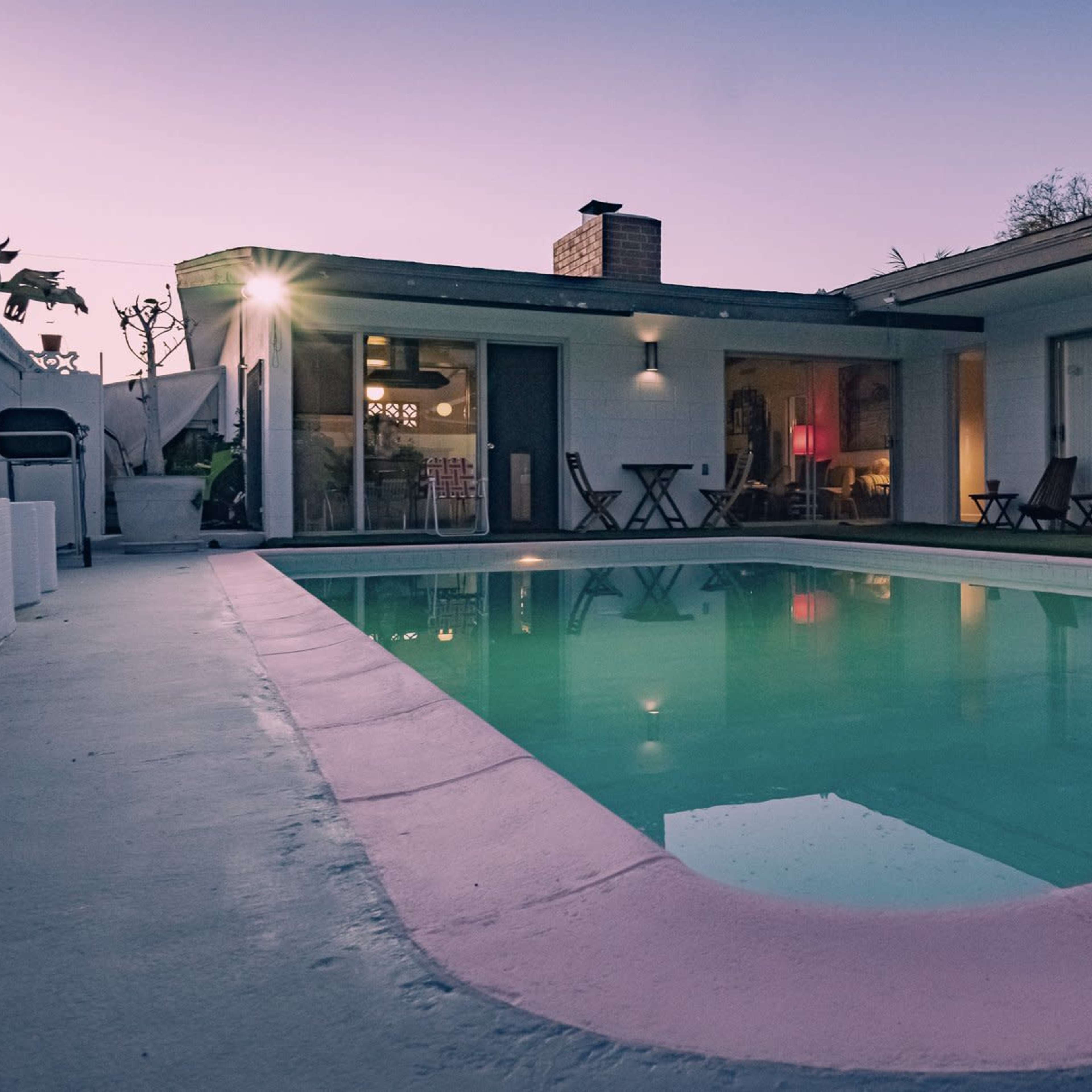 A swimming pool surrounded by a patio and a modern house with large windows at dusk.