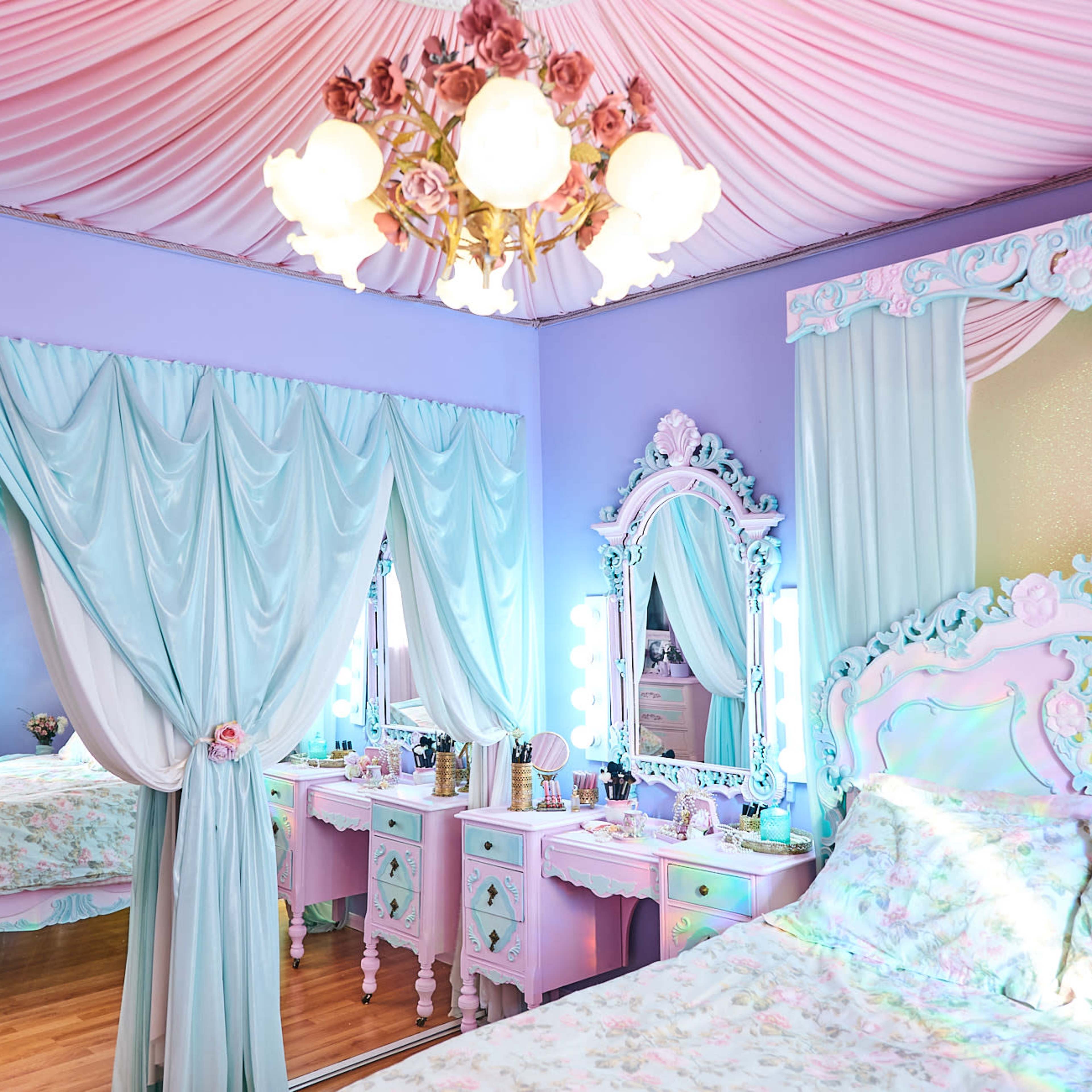 A pastel-colored bedroom with a floral-themed bed, draped curtains, and a vintage-style mirror and vanity set.