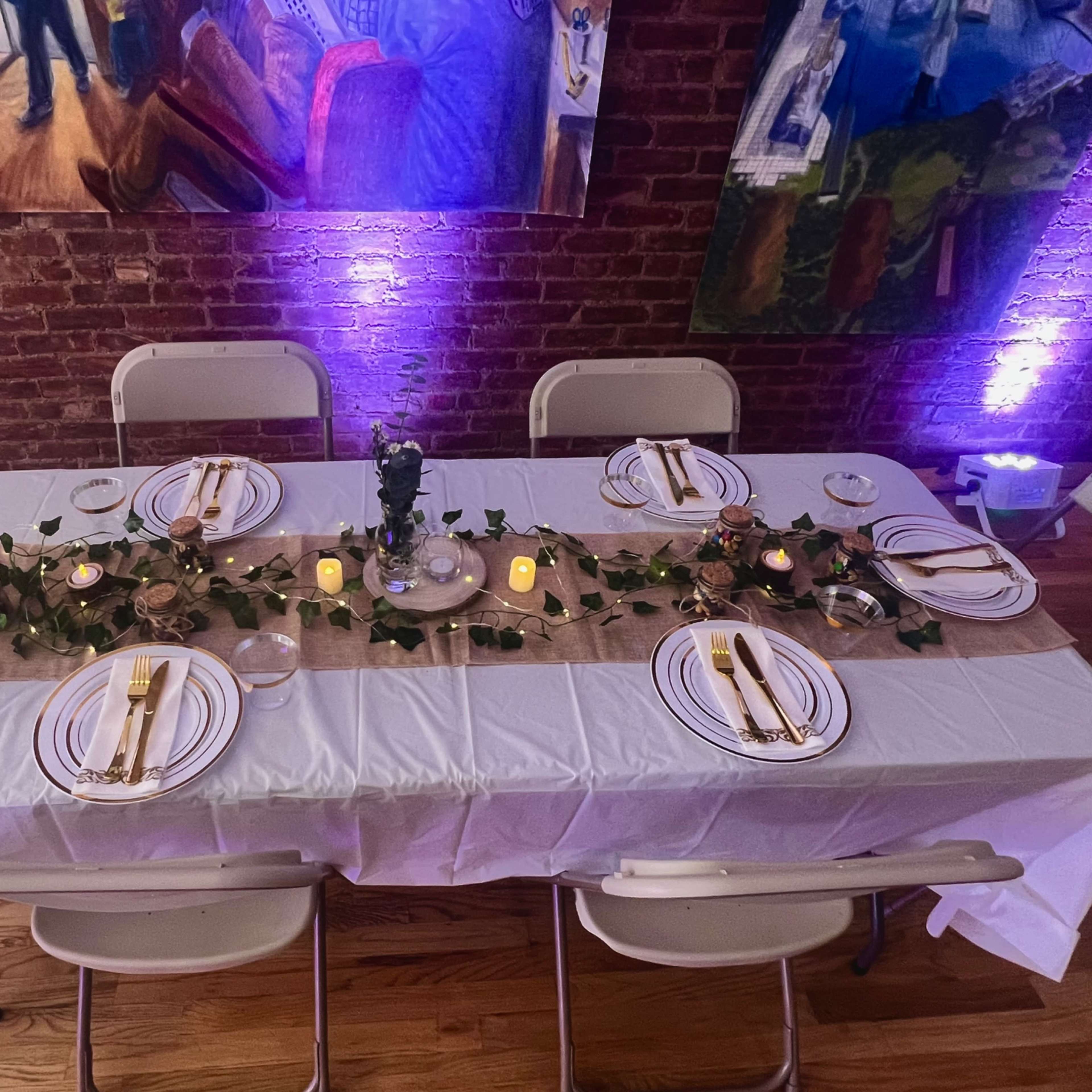 A dining table is set with white tablecloths, decorative plates, utensils, candles, and greenery, surrounded by chairs in a warmly lit room with brick walls and artwork.