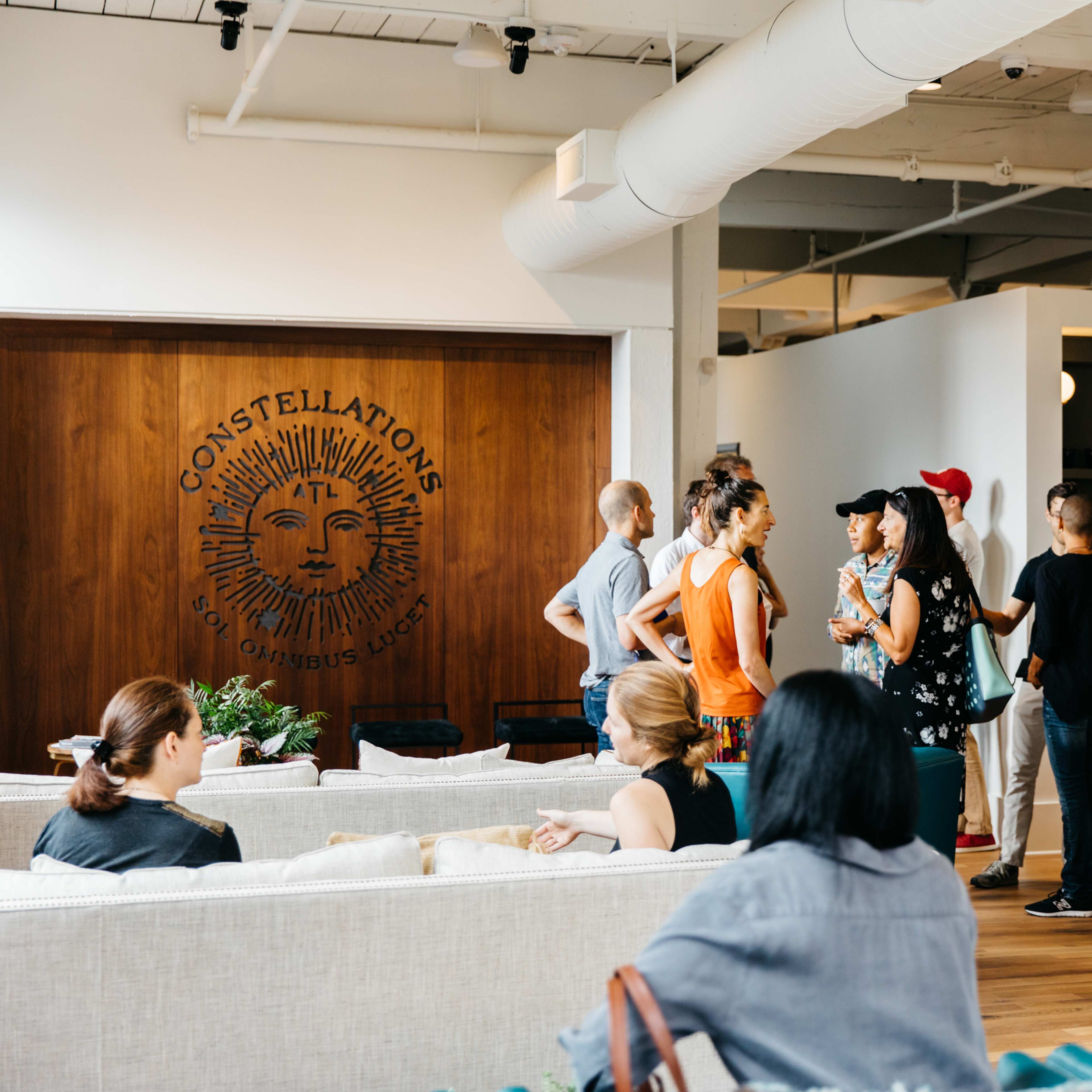 A group of people is gathered in a modern, open space featuring a decorative wall with the word "CONSTELLATIONS" and various seating arrangements.