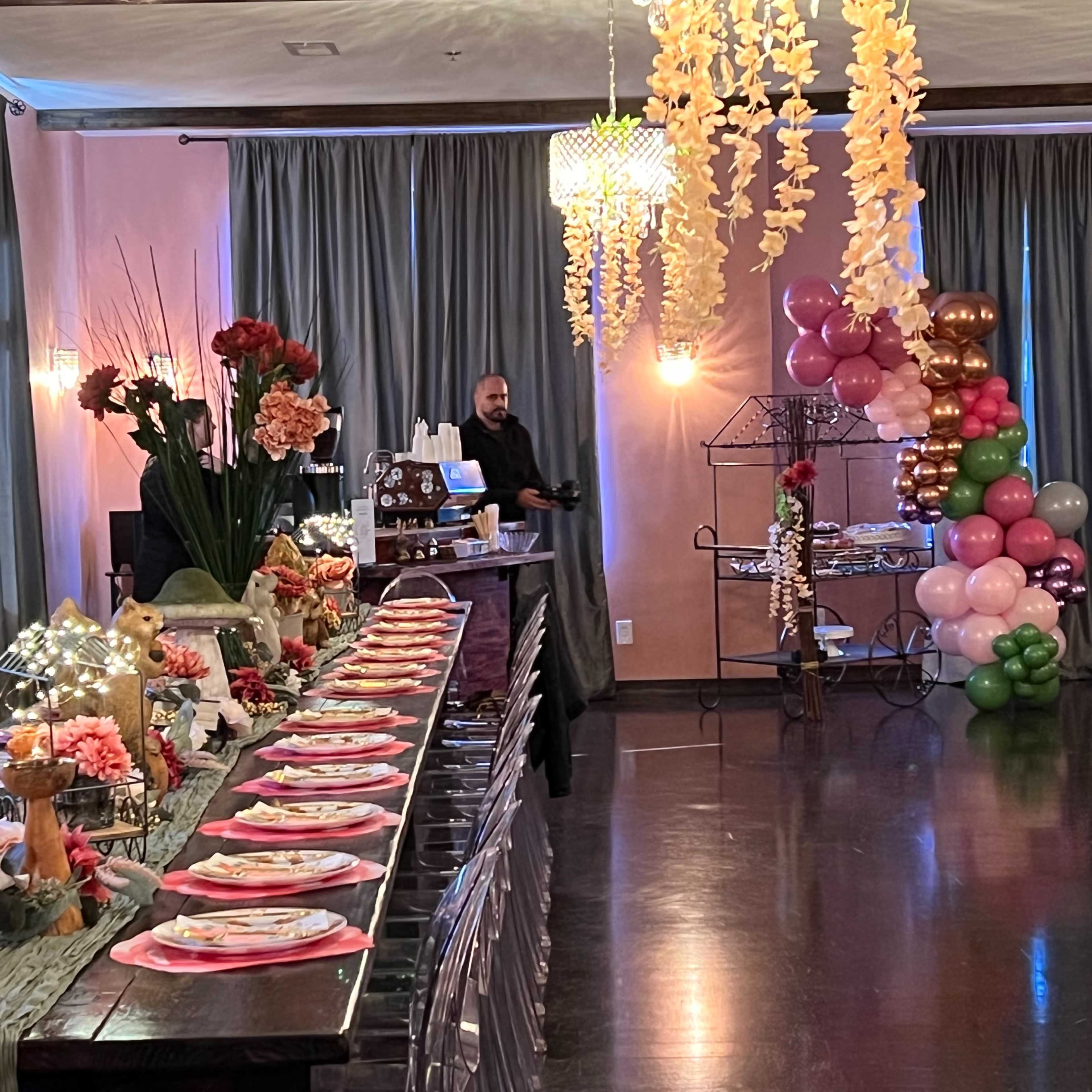 A decorated event space with a long dining table set with floral arrangements, plates, and a dessert display, while a man stands behind a counter near a balloon arrangement in the corner.