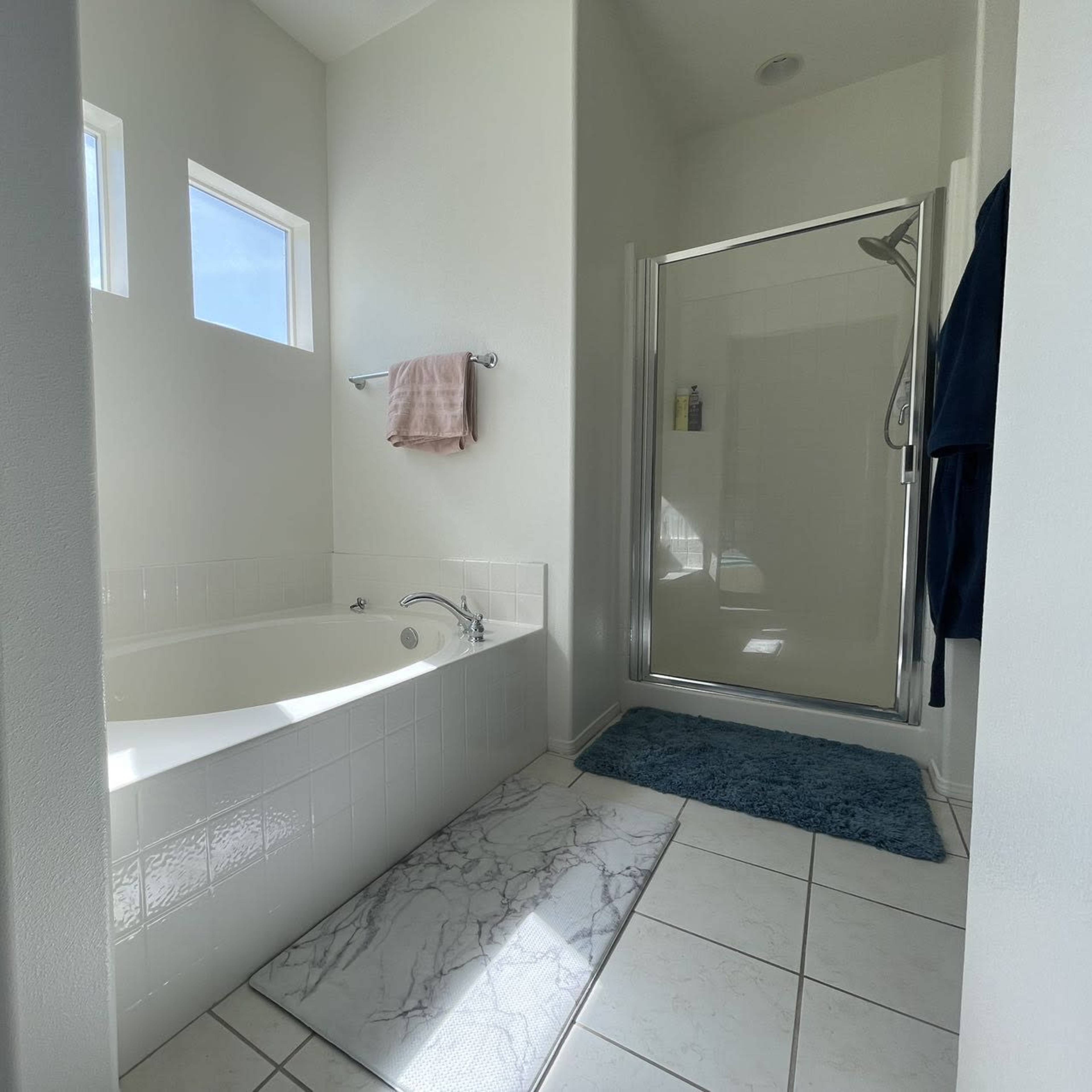 The bathroom features a bathtub next to a glass-enclosed shower, with two small windows and light-colored walls.
