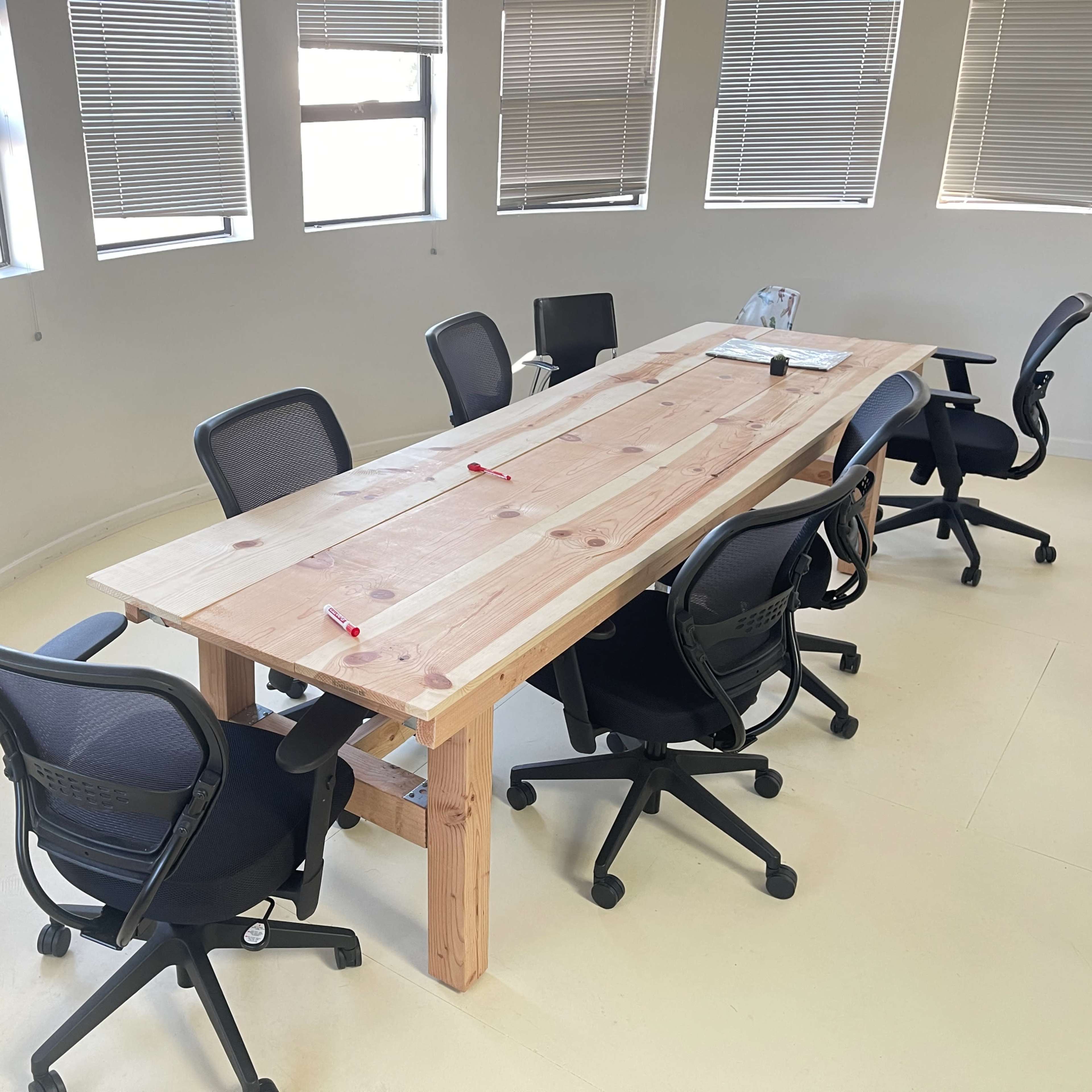 A long wooden table surrounded by black office chairs is located in a well-lit meeting room with large windows.