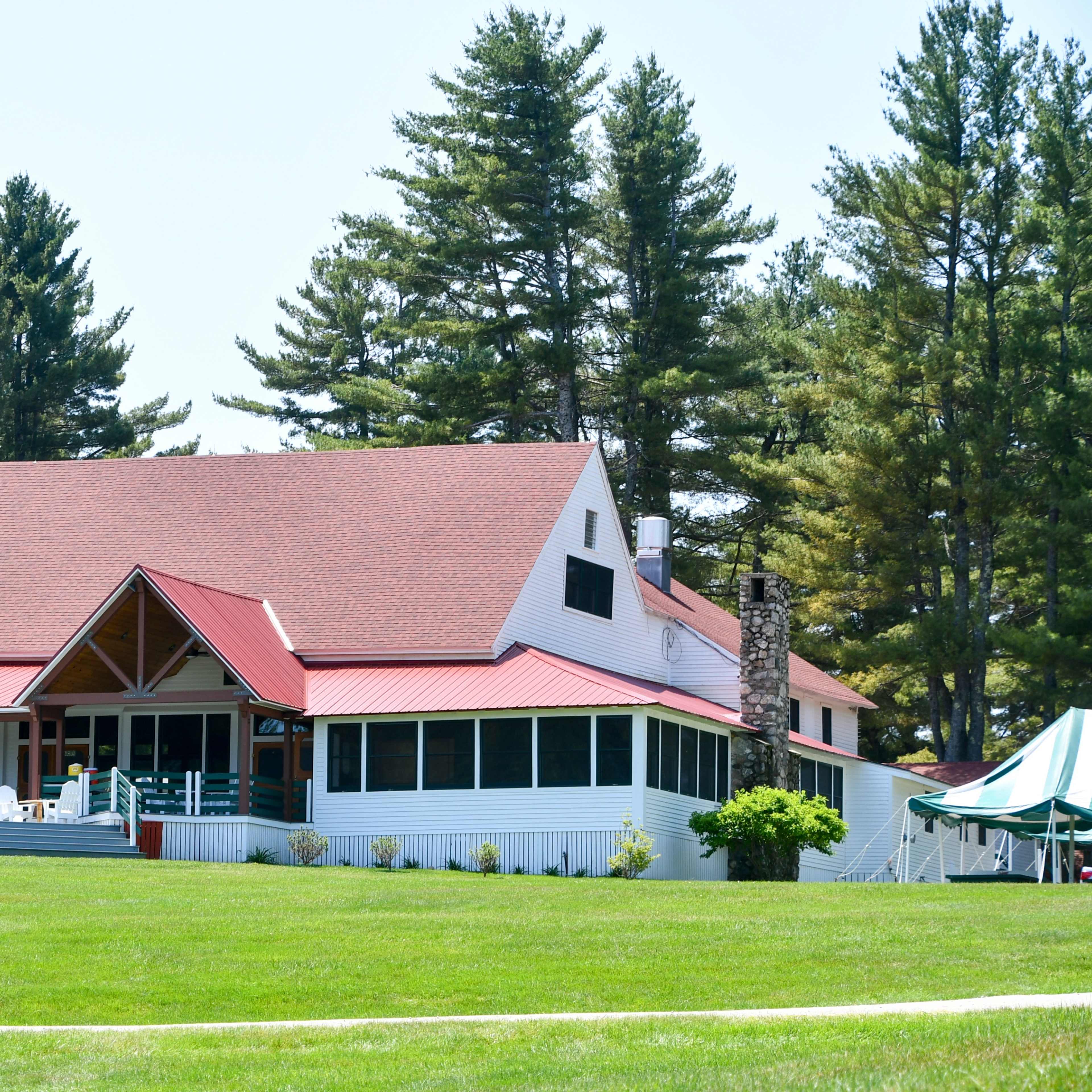 A large white house with a red roof and multiple porches is set against a backdrop of tall pine trees.