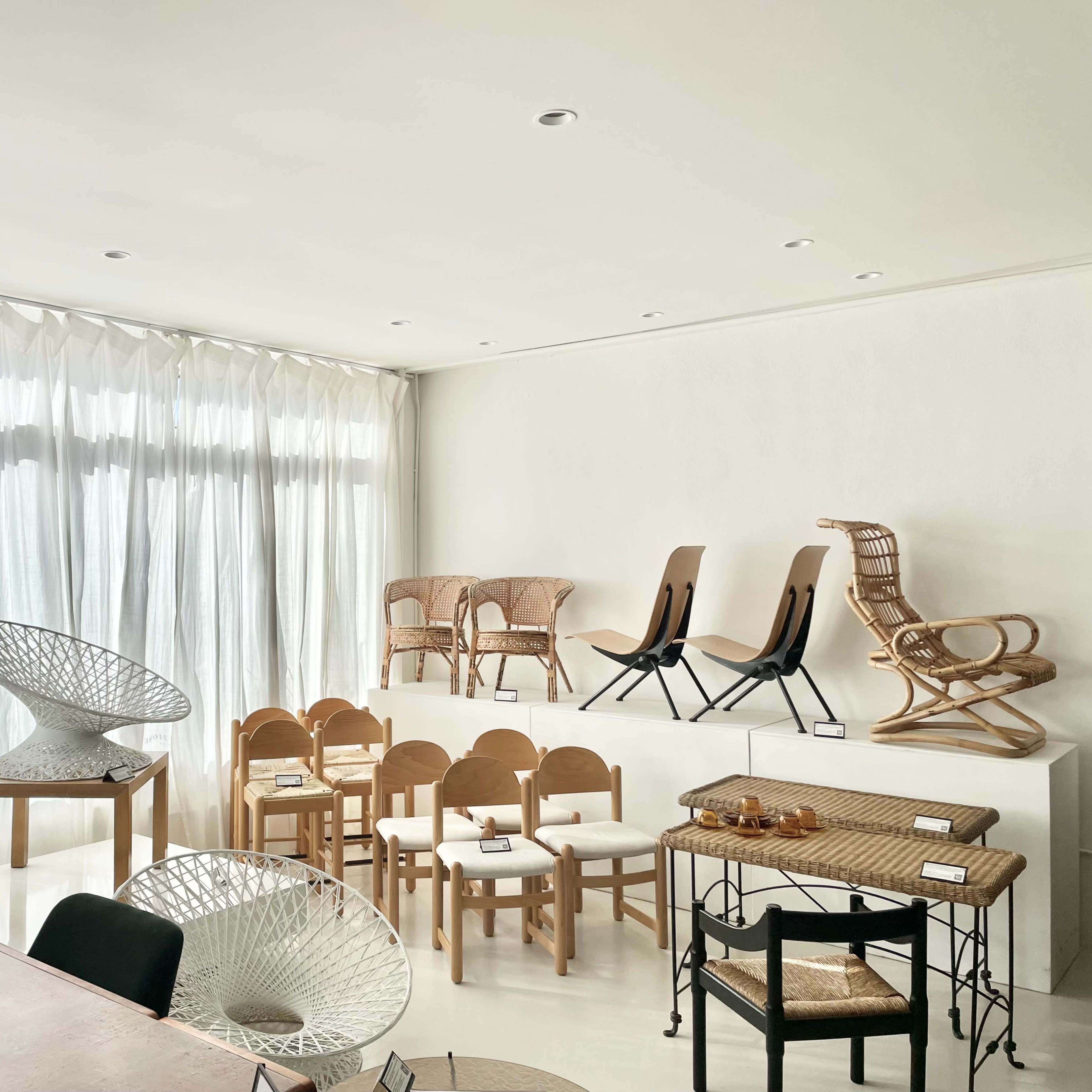 A modern, minimalist interior space featuring various wooden and wicker chairs, a round sculpture, and large windows with sheer curtains.