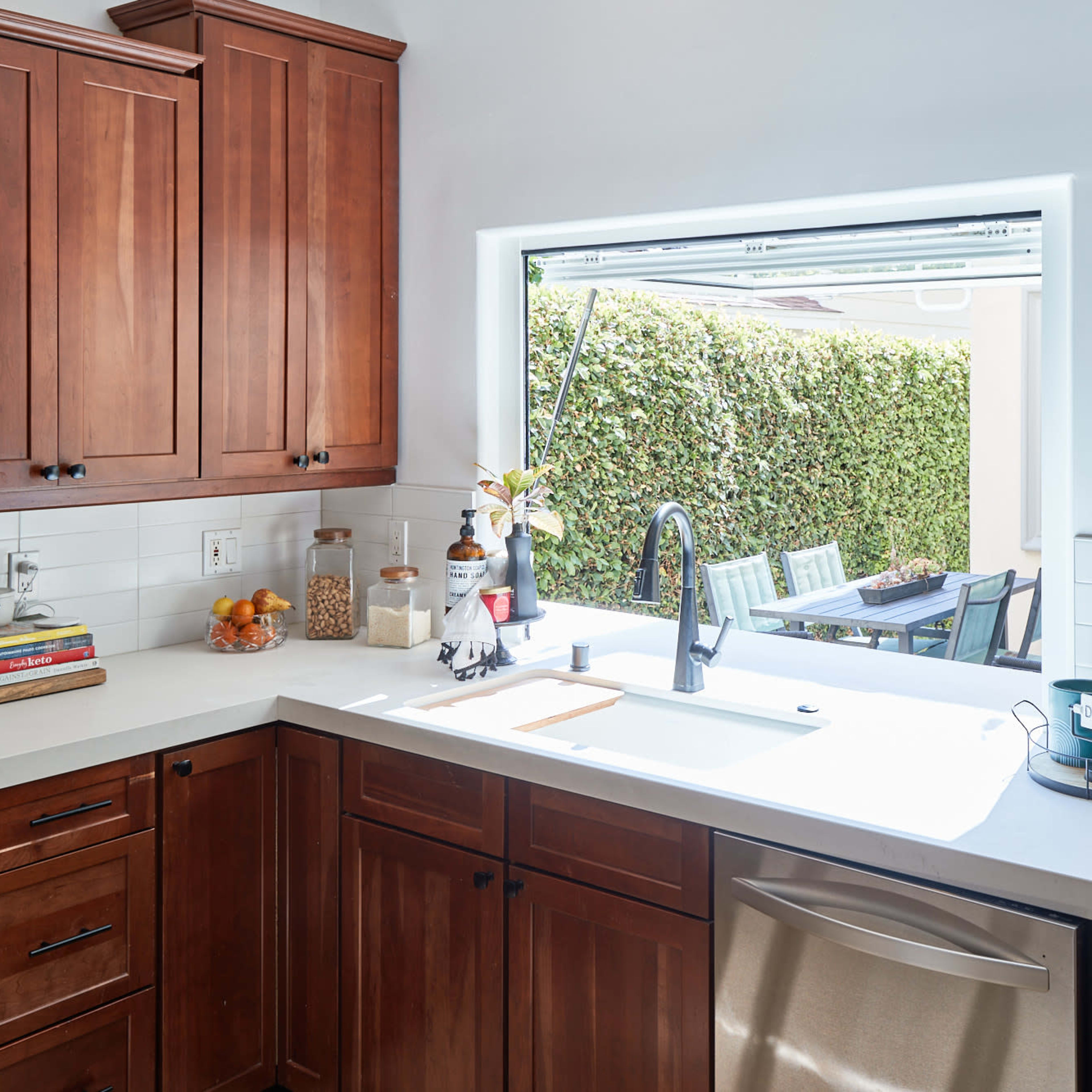 A kitchen with wooden cabinets and a white countertop features a window that opens to a backyard with green foliage.