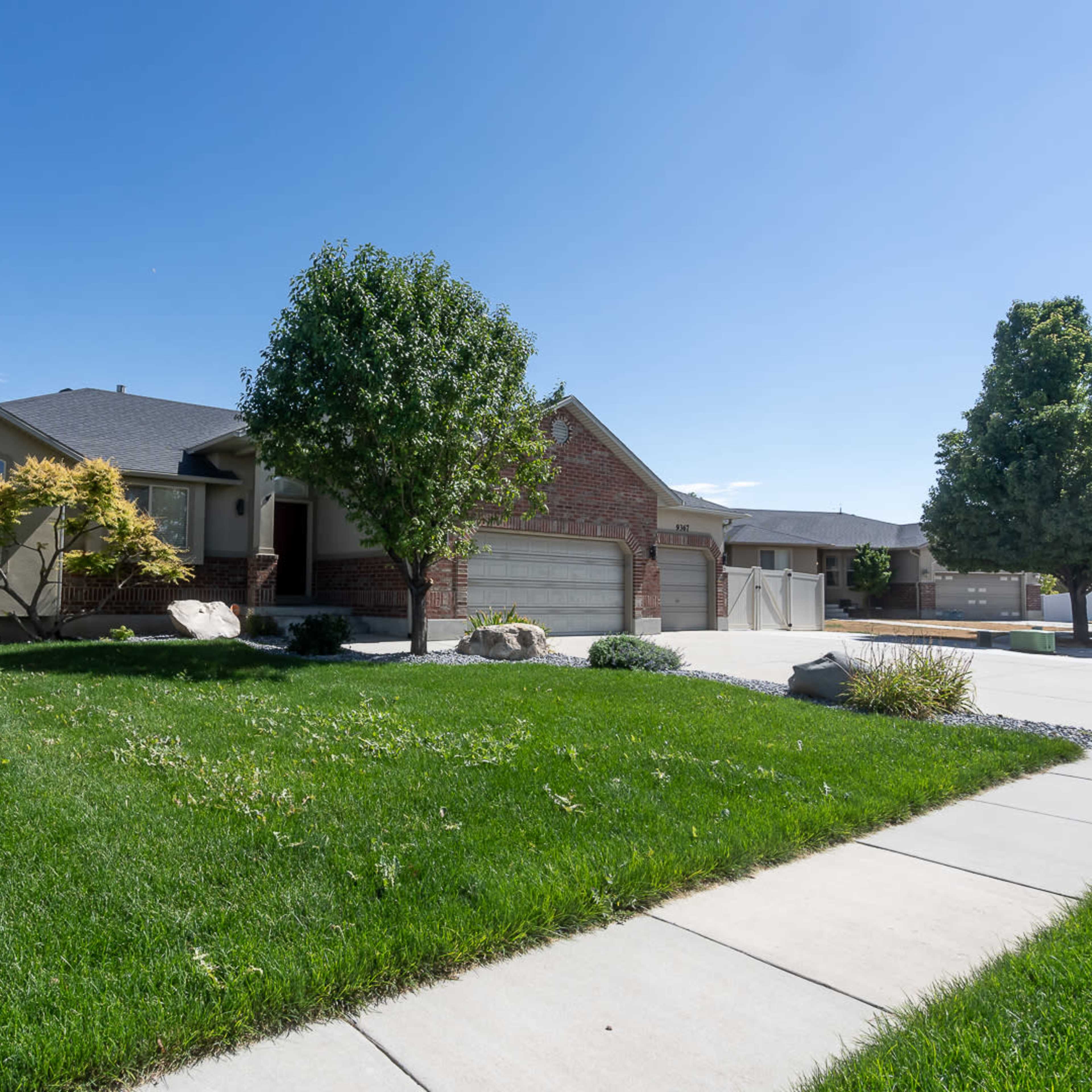 A single-story house with a brick facade and a landscaped yard featuring various trees and shrubs is seen under a clear blue sky.