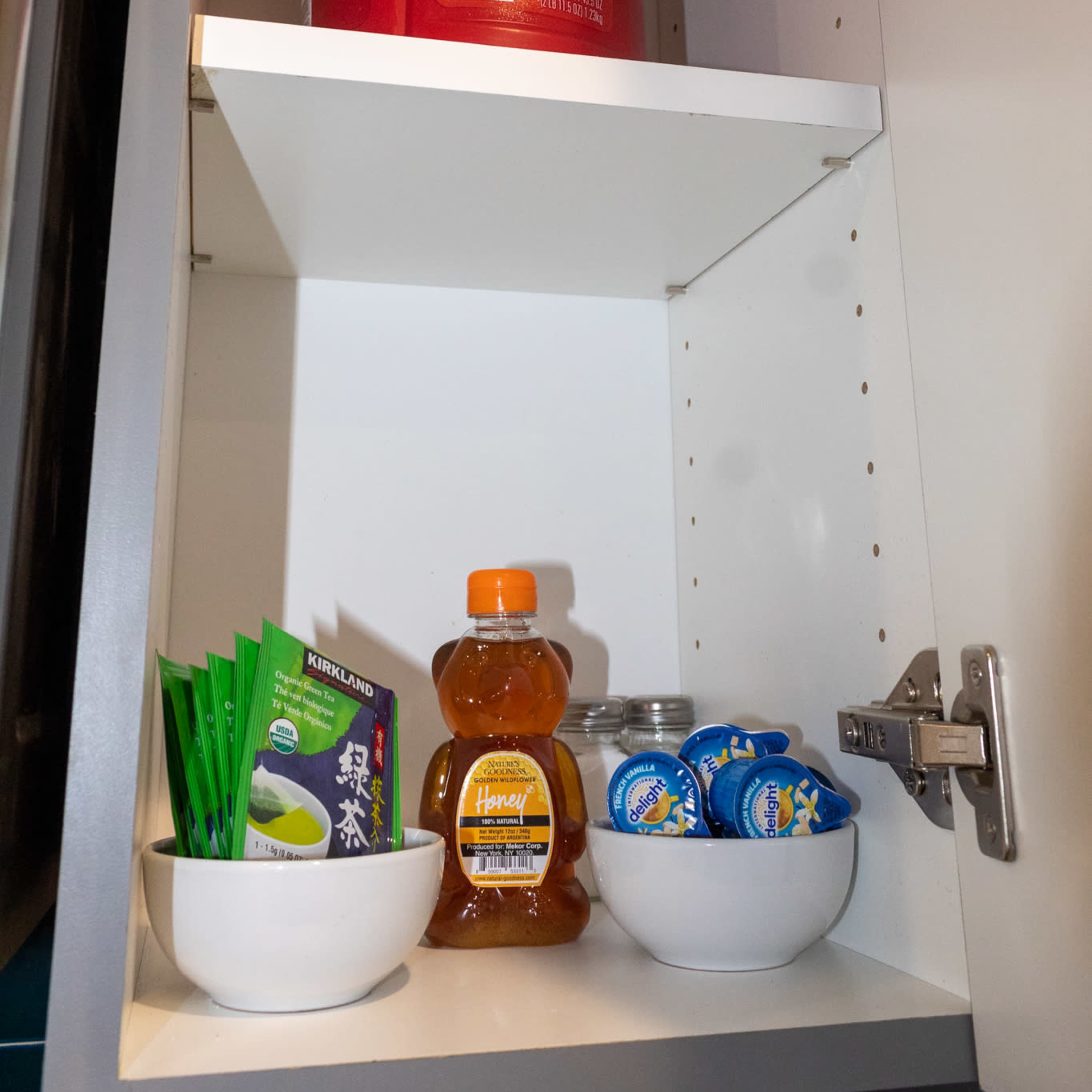 A kitchen cabinet shelf contains a large can of Folgers coffee, a bottle of honey, and several containers of butter, along with packets of tea.