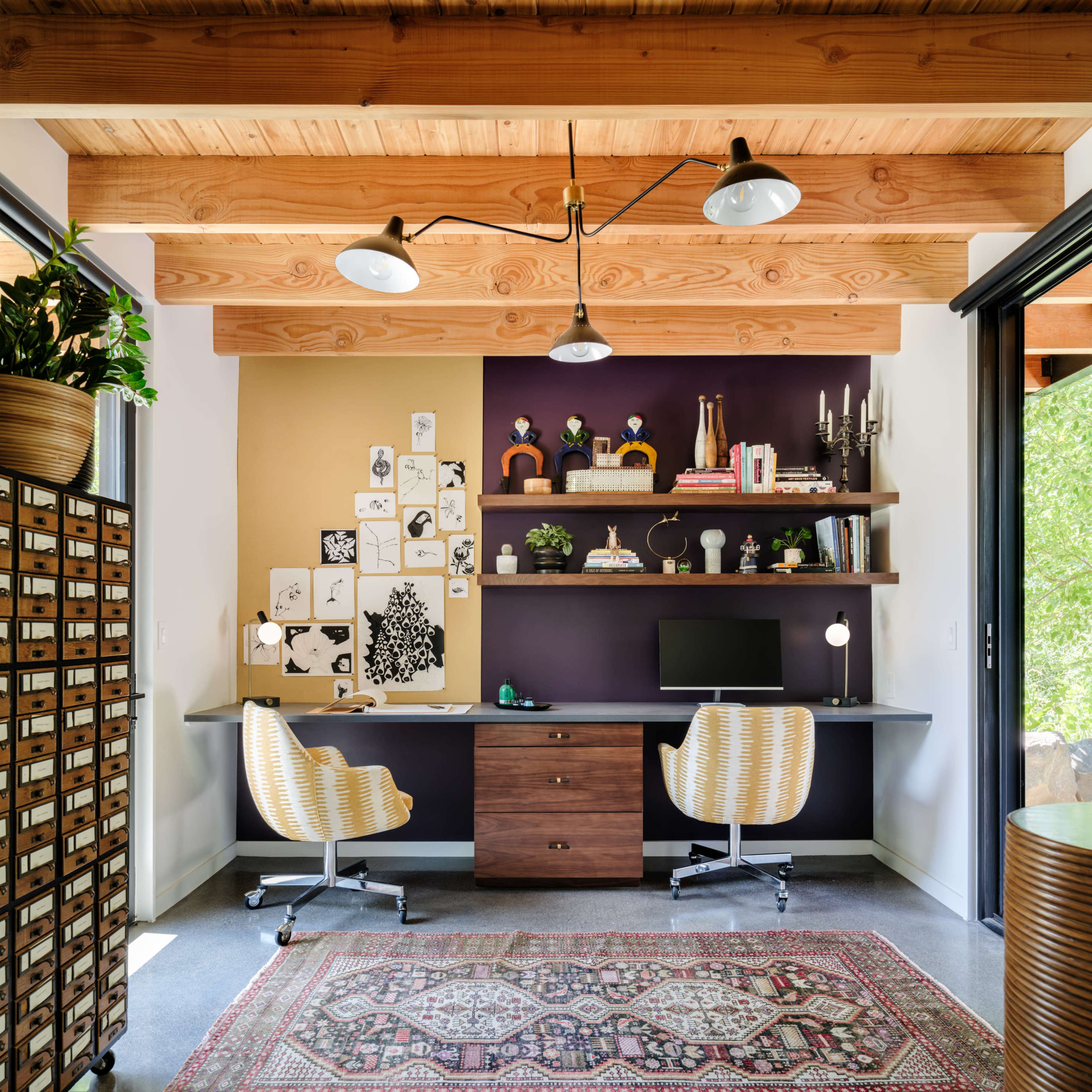 A modern home office with a wooden ceiling, a desk against a purple accent wall, shelving, and a vintage rug.