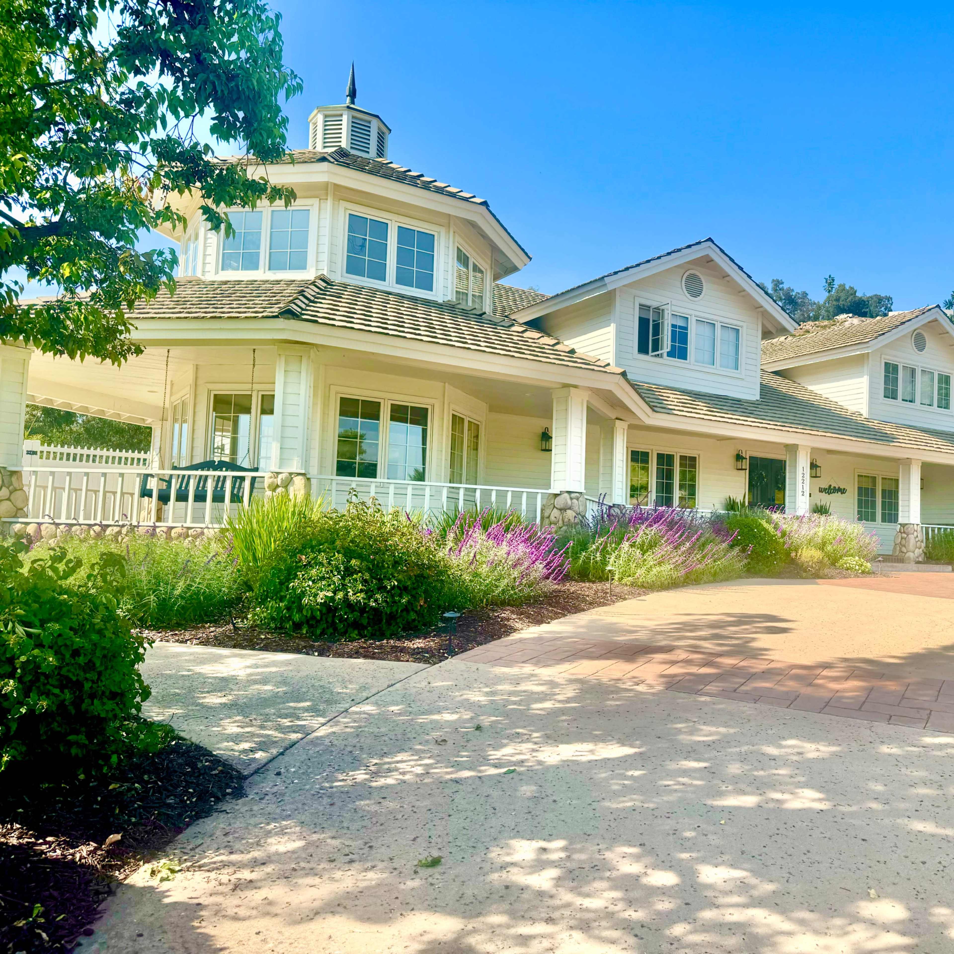 A large white house with a distinctive cupola is surrounded by colorful landscaping and a paved driveway.