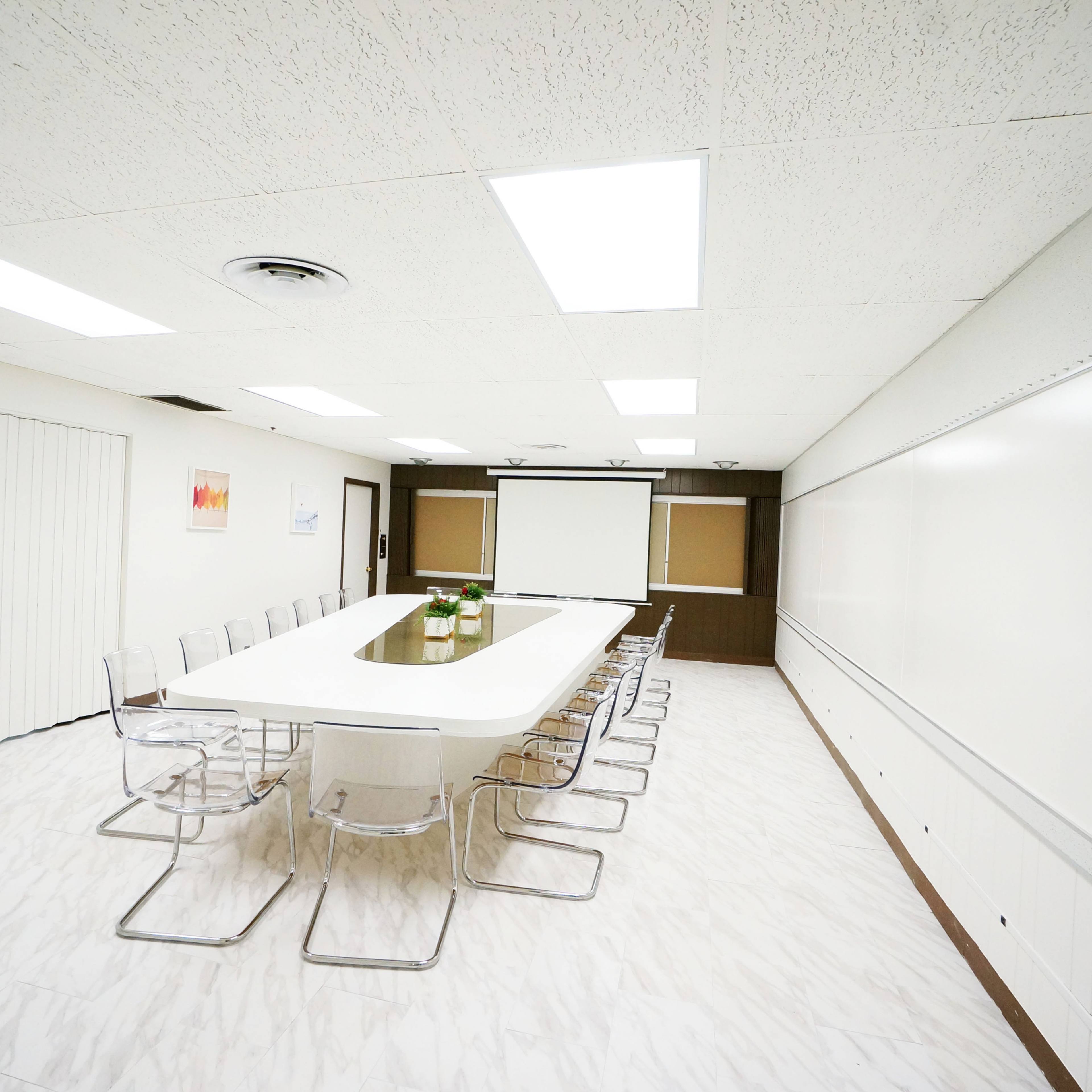A large conference room features a long white table surrounded by clear chairs, with a projector screen and whiteboard on one wall.