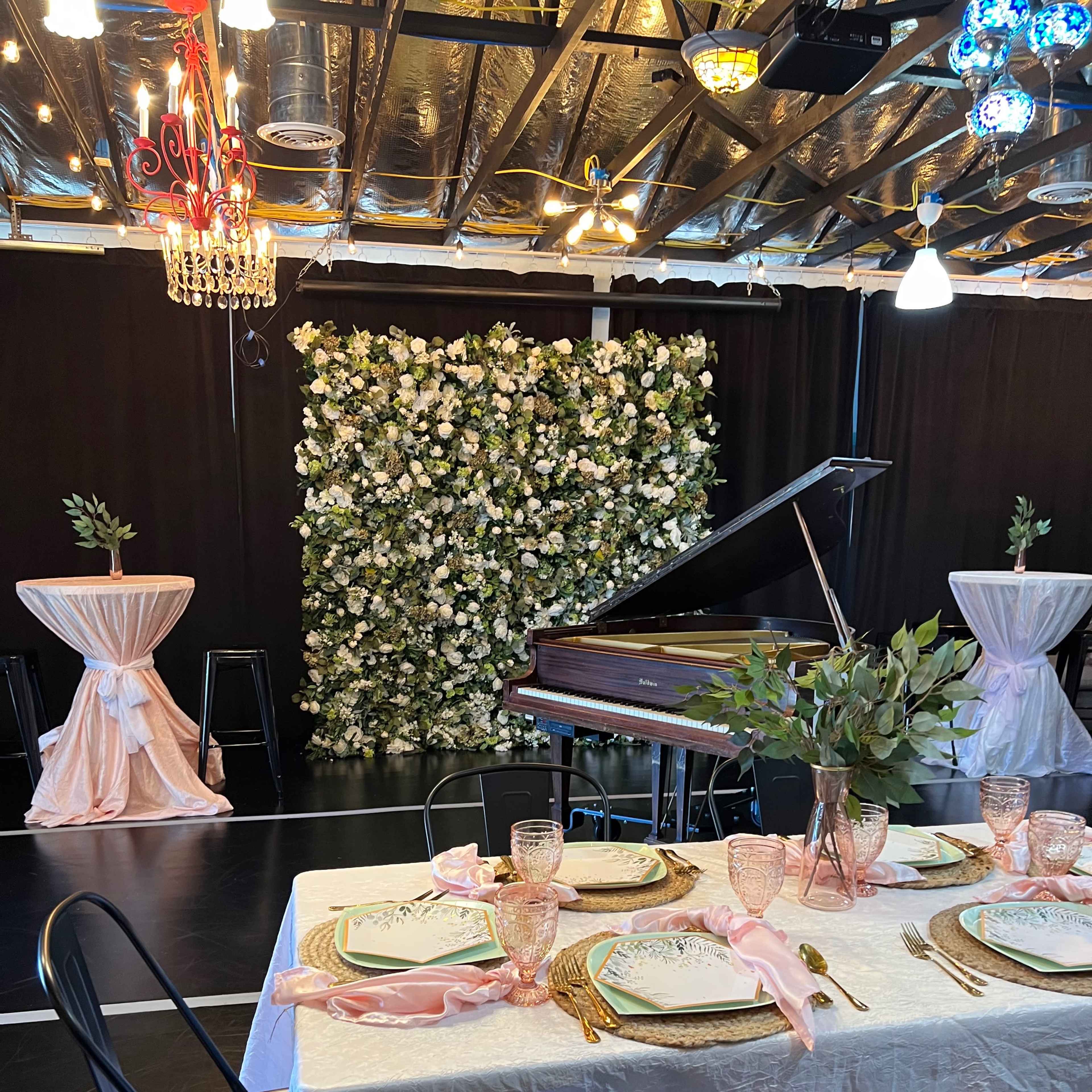 A decorated event space features a grand piano in front of a floral backdrop, with a dining table set for a gathering and tall cocktail tables nearby.