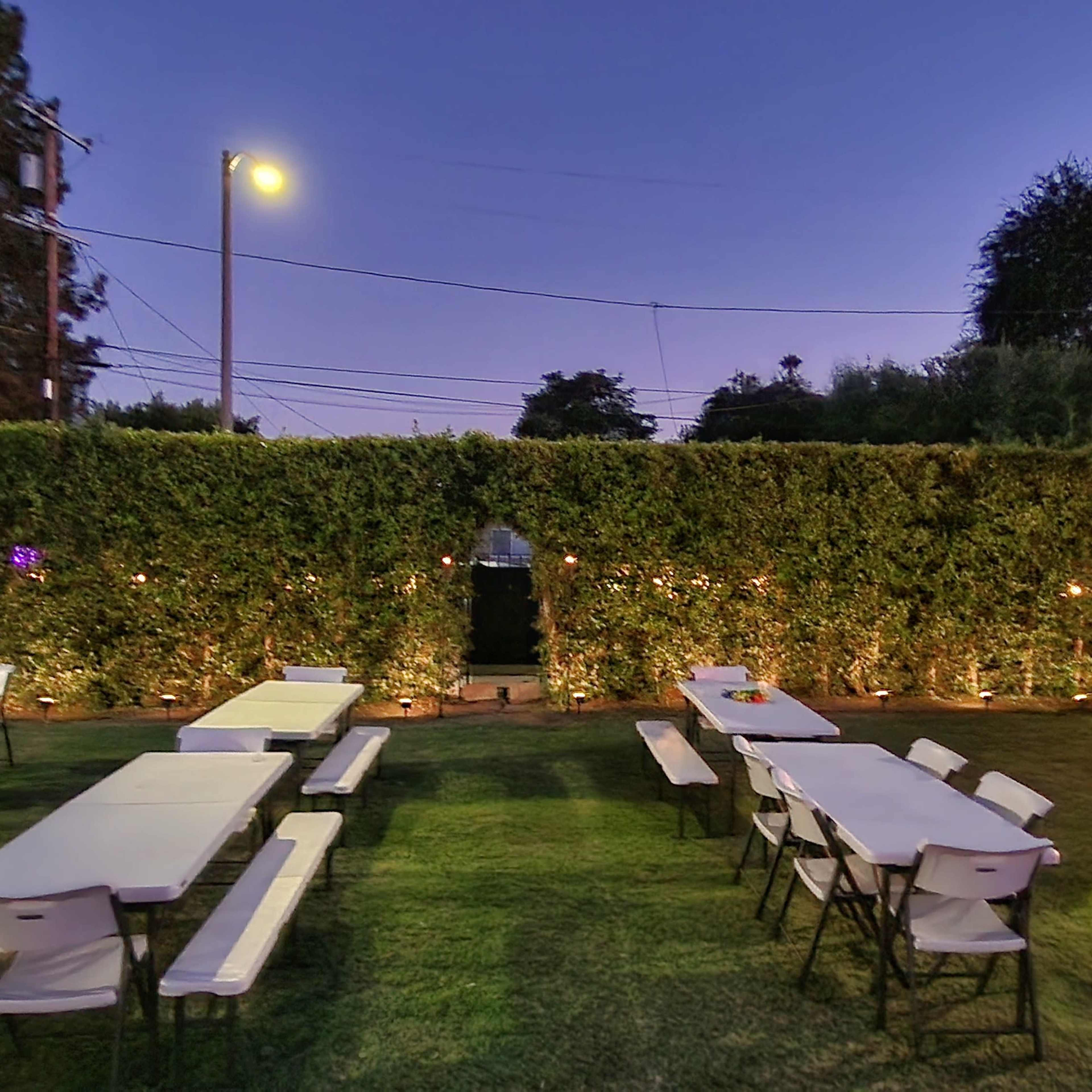 A backyard setting with several white tables and benches arranged on a green lawn, surrounded by a leafy hedge and illuminated by evening lights.