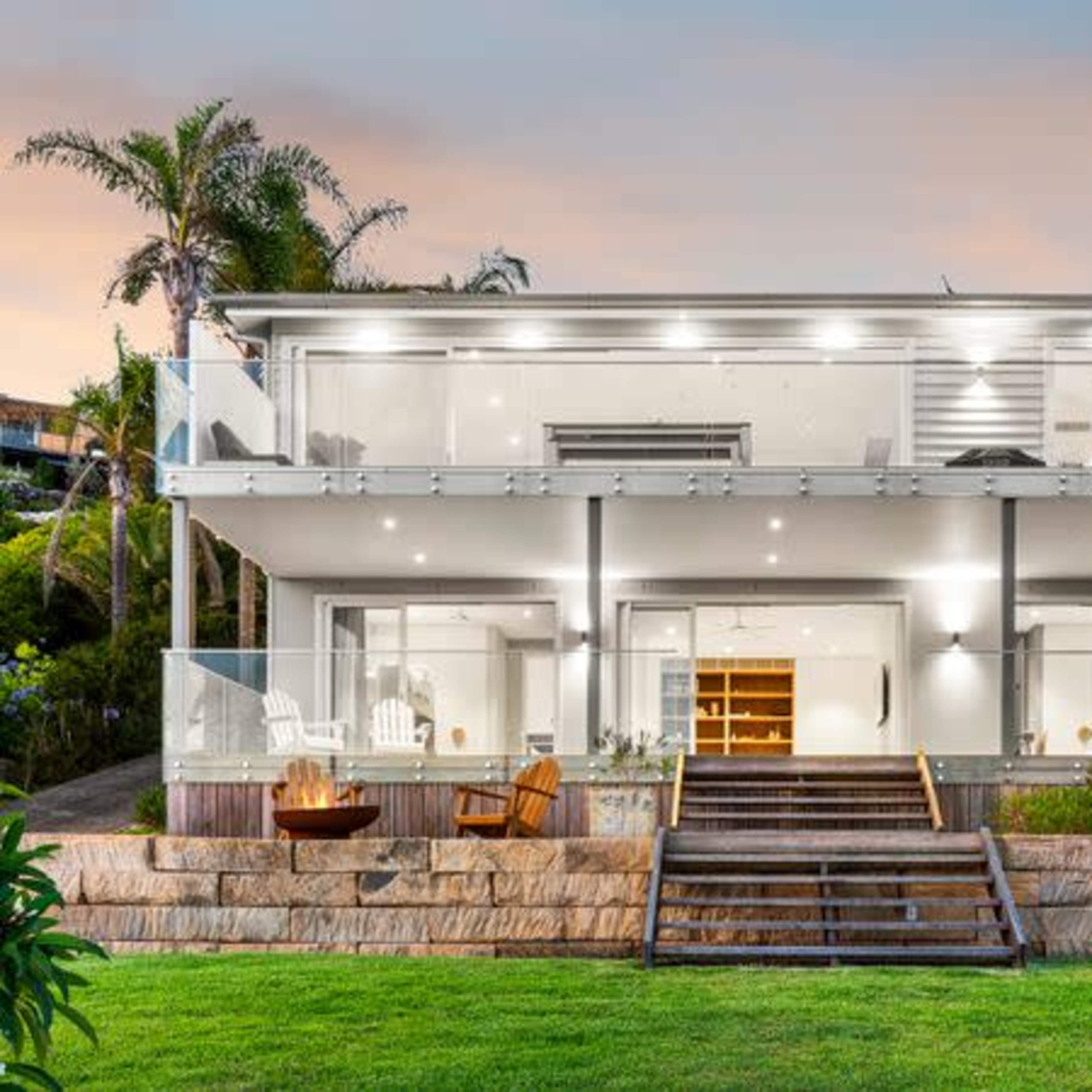 A modern two-story house with large glass balconies, wooden steps leading to a grassy yard, and palm trees in the background.