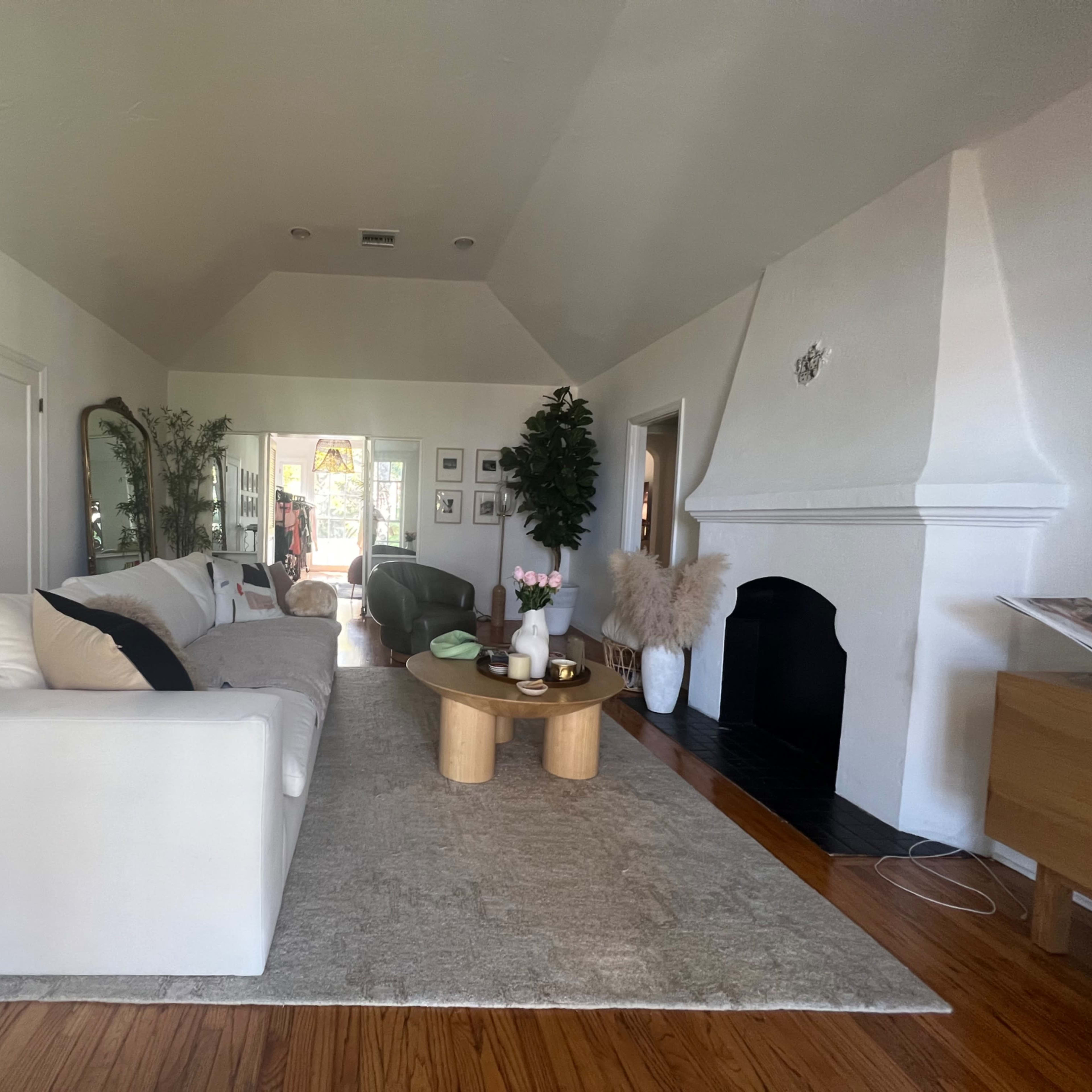 A bright living room with a white sofa, a wooden coffee table, and a large window leading to an outdoor area.
