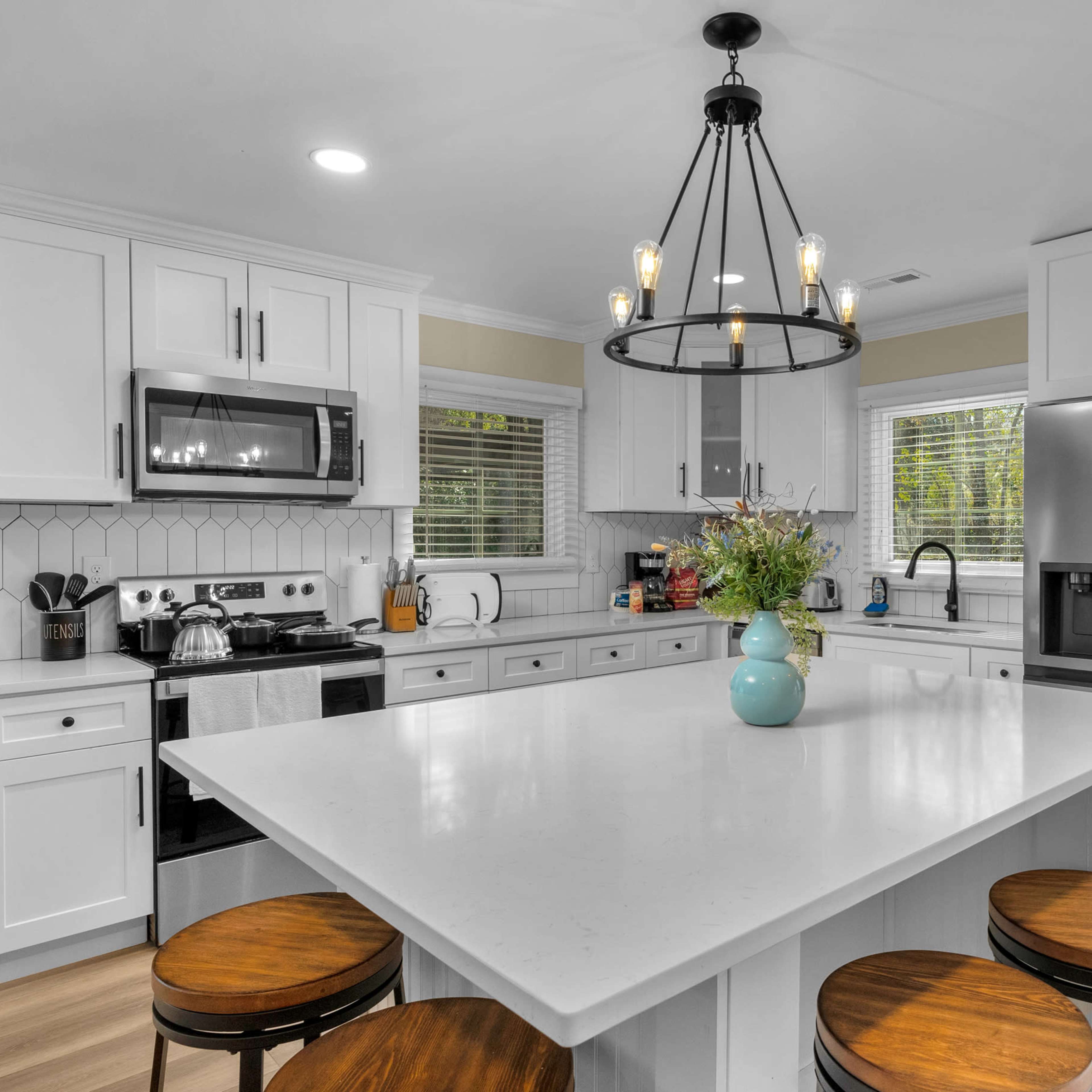 A modern kitchen features a central island with four wooden stools, stainless steel appliances, and ample natural light from two windows.
