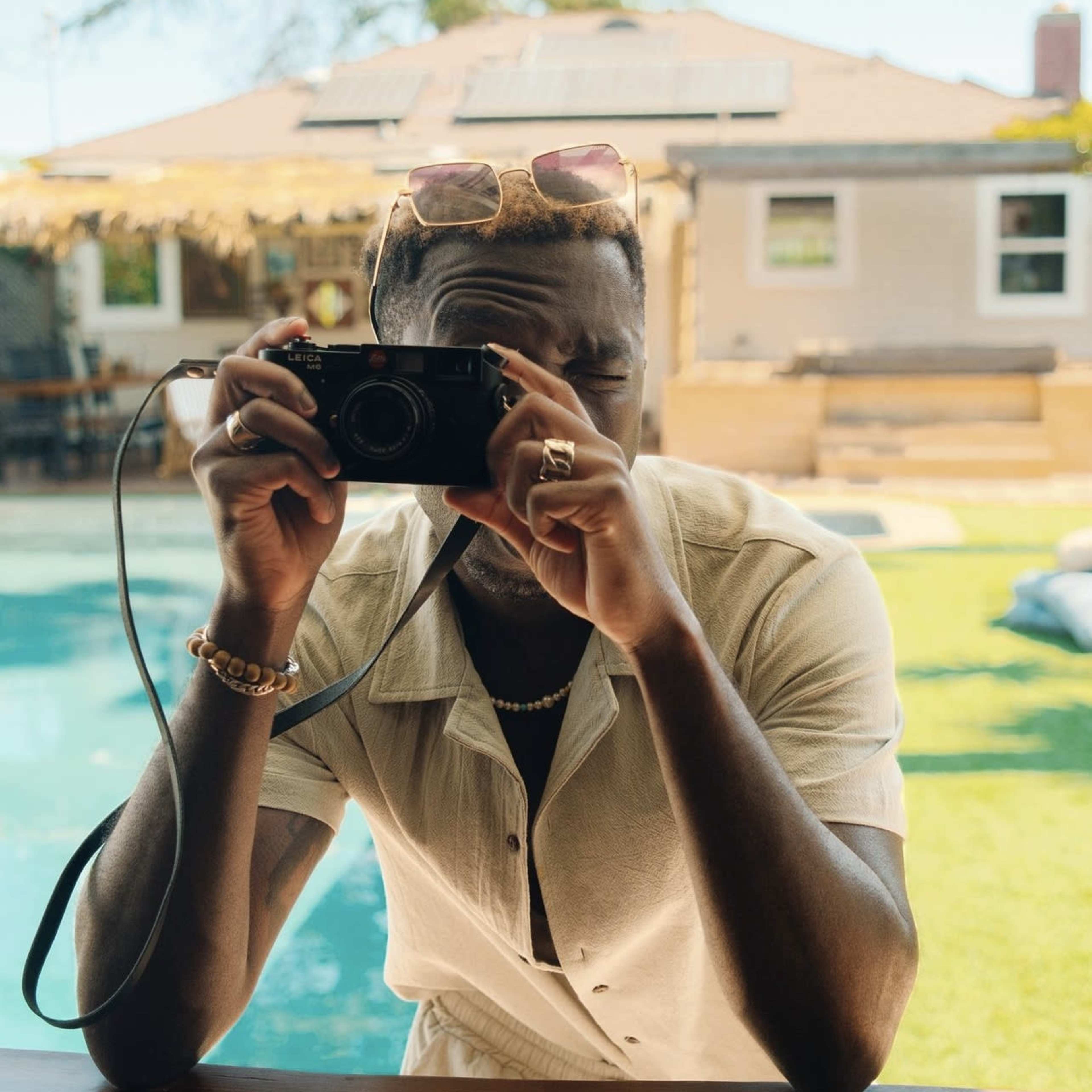 A person in a light-colored shirt holds a camera up to their face while standing near a pool, with a house and trees visible in the background.