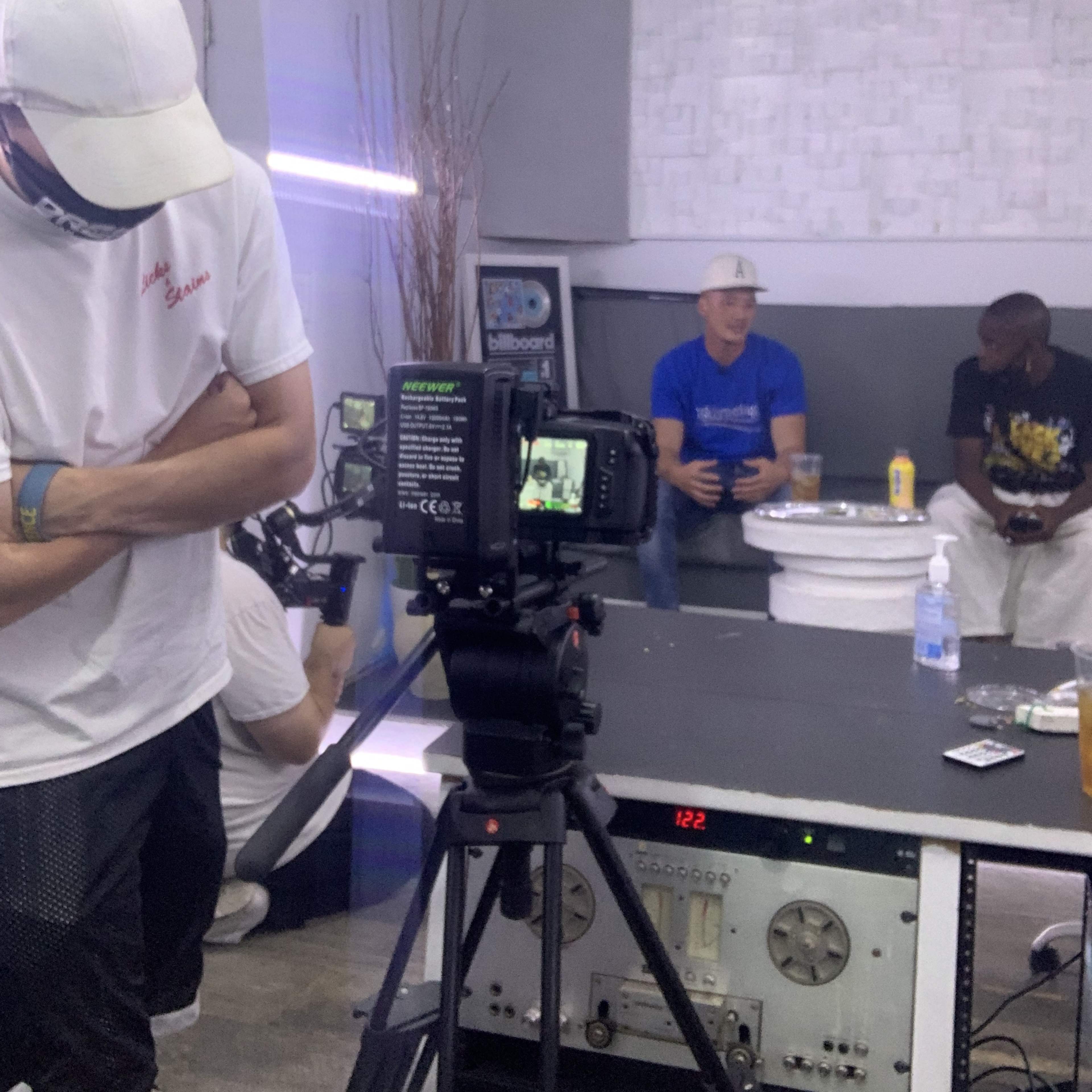 A person wearing a white cap stands in the foreground while two individuals sit in the background, engaged in conversation in a recording studio.