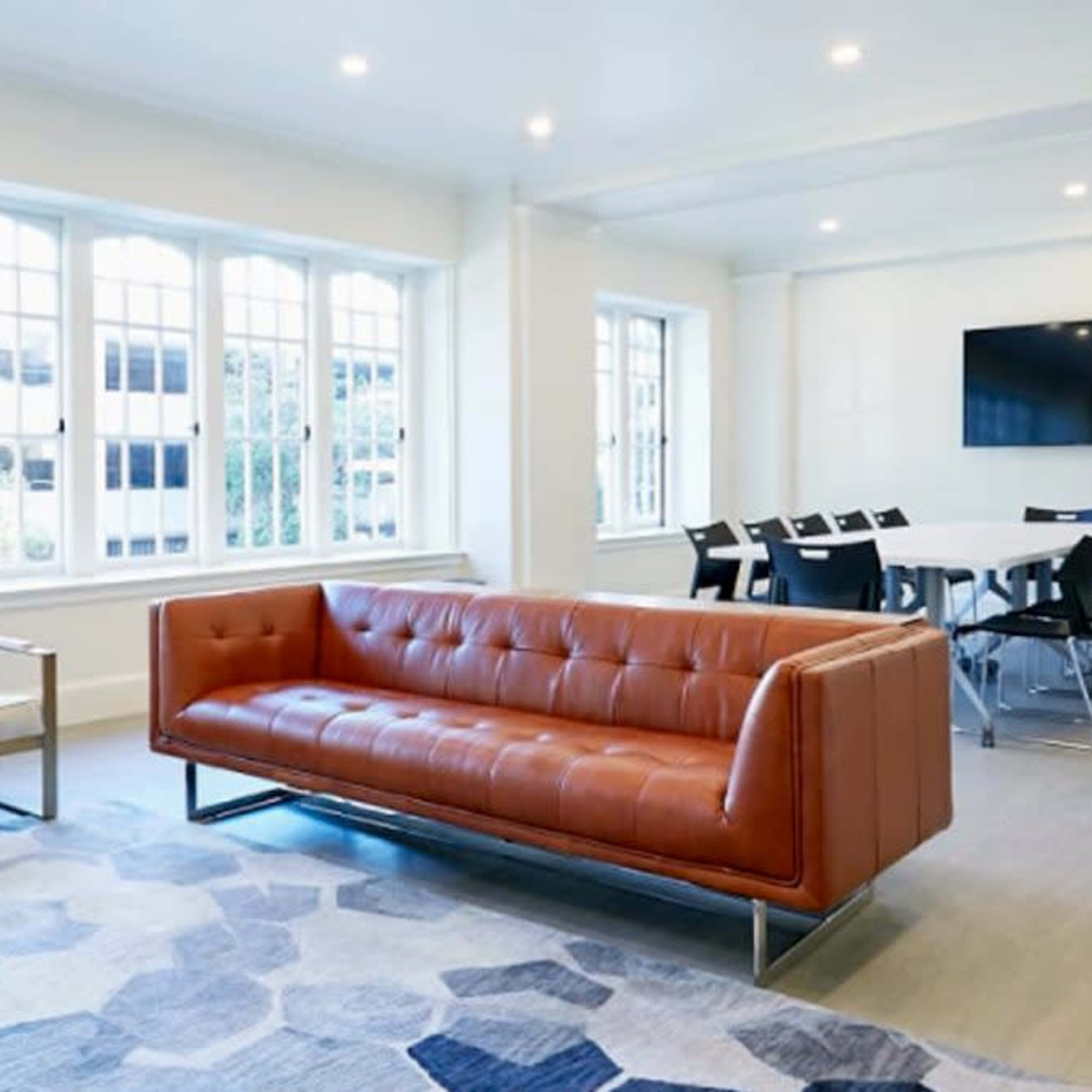 A modern office space featuring a brown leather sofa, two armchairs, a large rug with a geometric pattern, and a conference table surrounded by black chairs, all in a well-lit room with multiple windows.