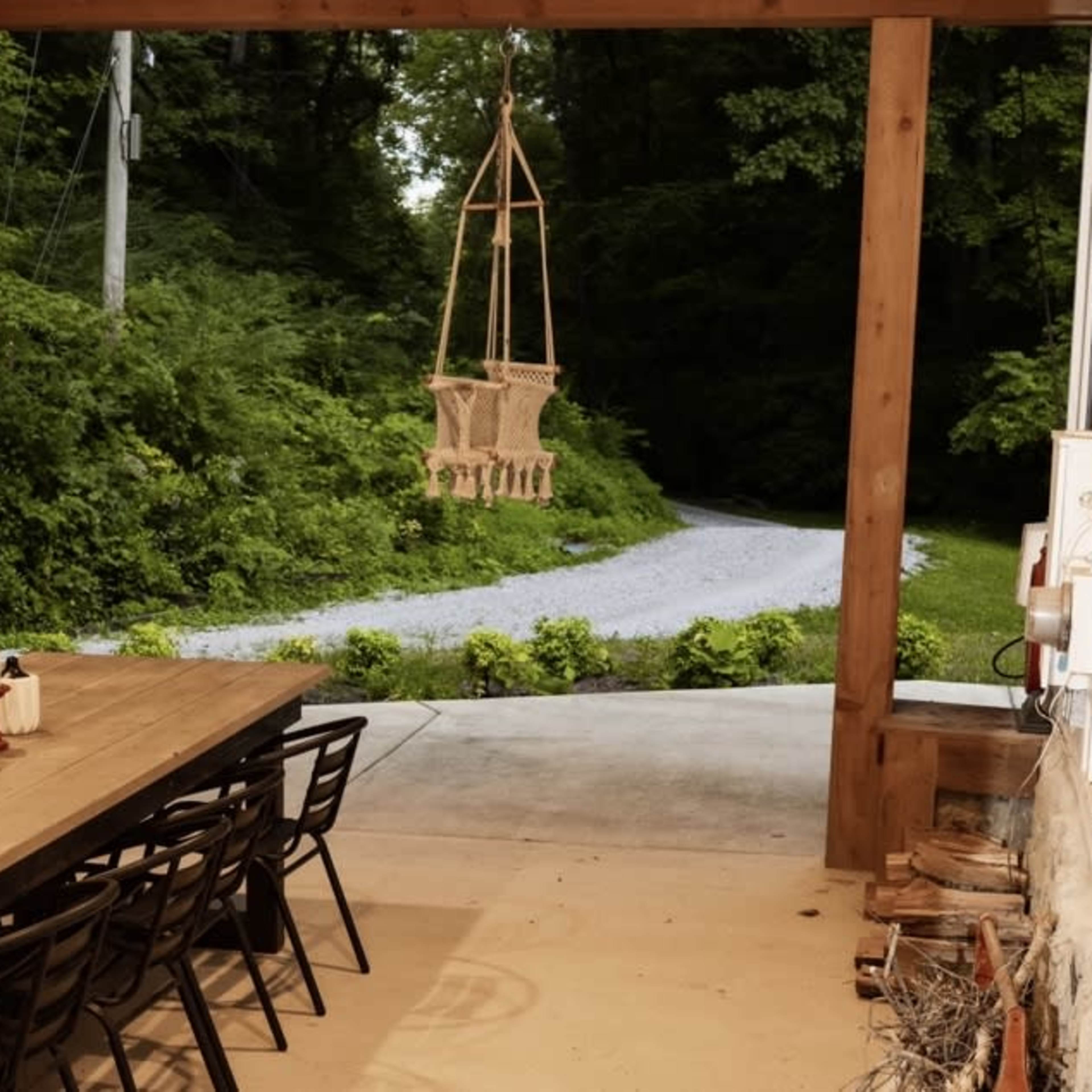 A covered outdoor area with a long wooden dining table and a hanging chair, overlooking a gravel pathway surrounded by greenery.