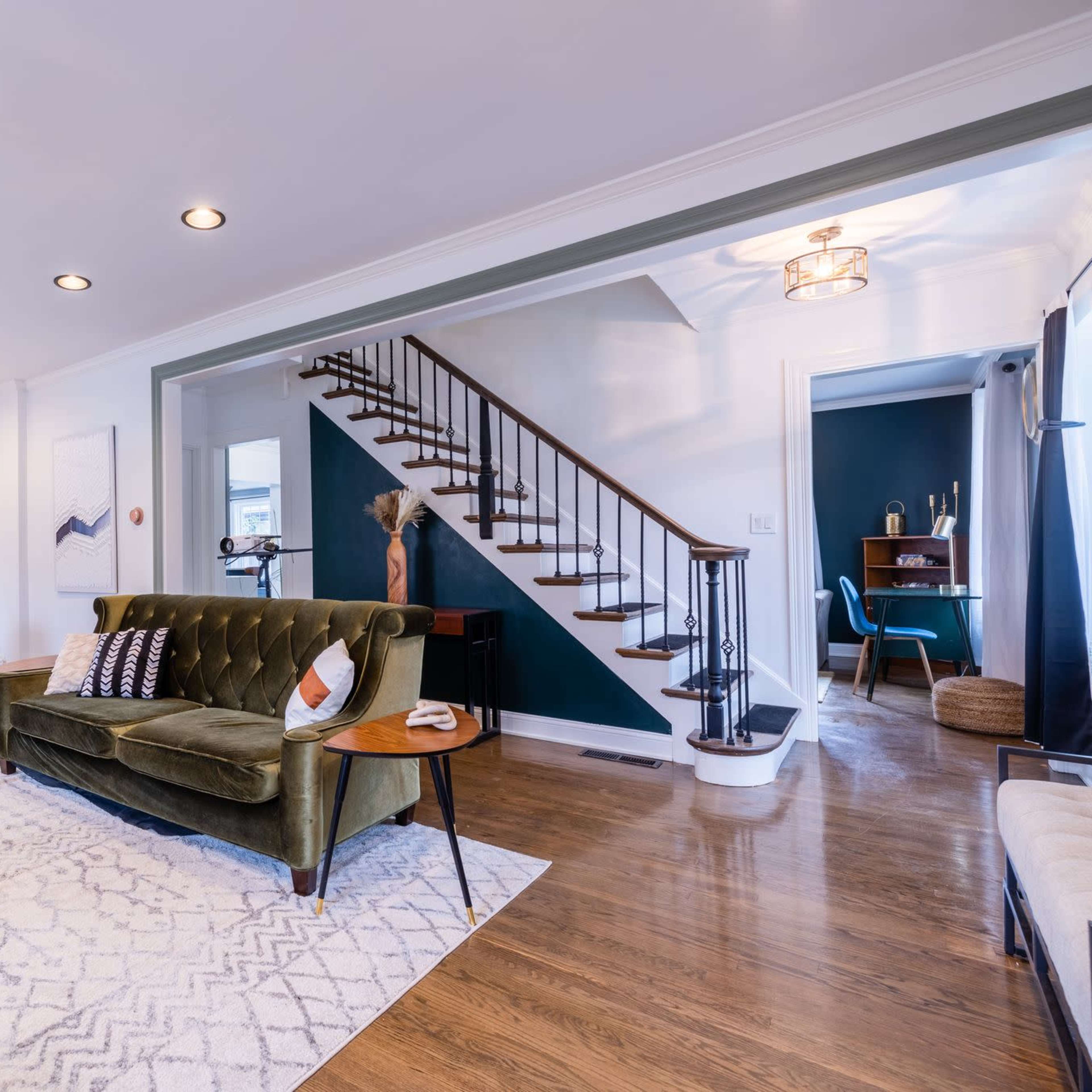A stylish living room with two green velvet sofas, a glass coffee table, and a staircase leading to an upper level.
