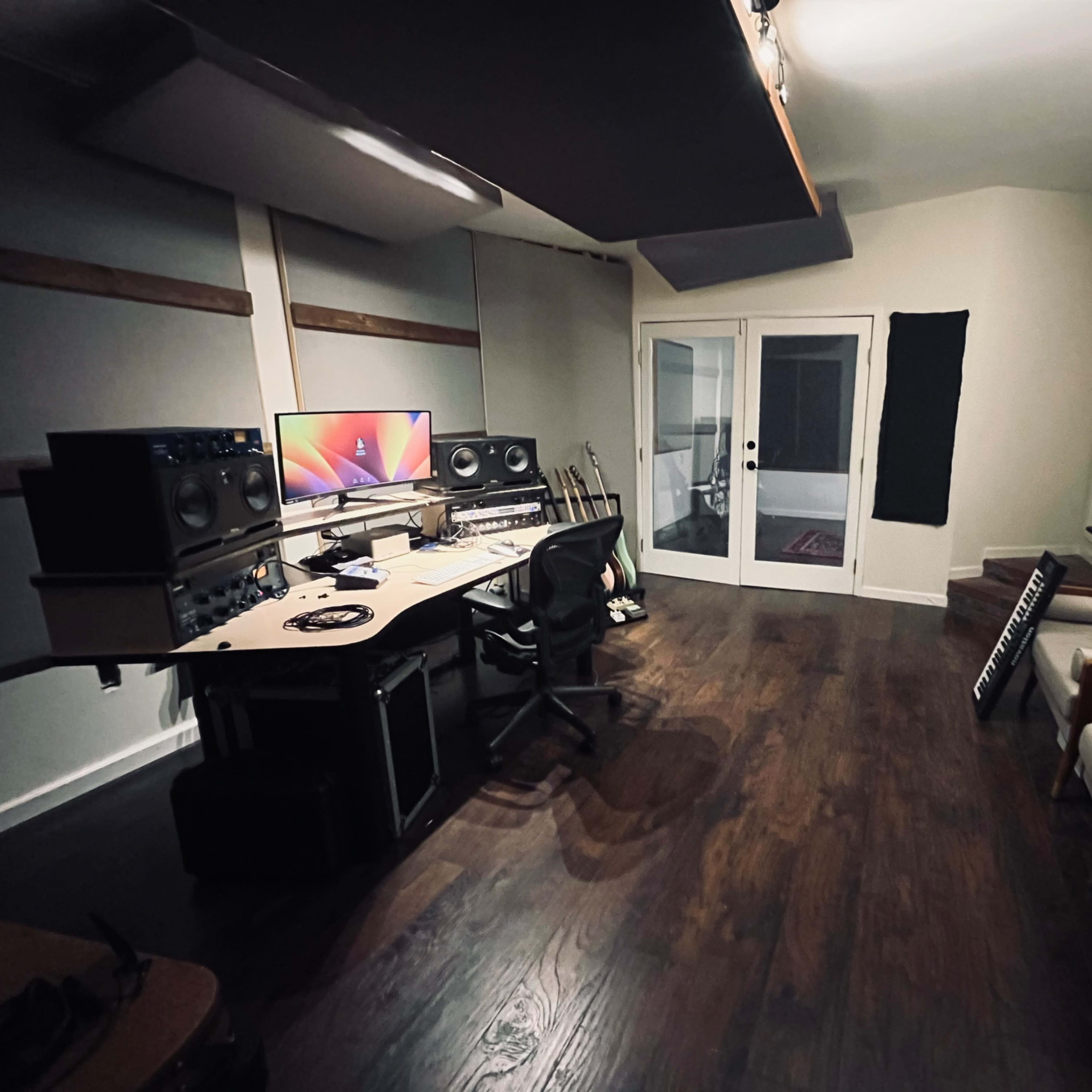 A music studio featuring a mixing desk with speakers and a computer setup, a sofa, and a glass door leading to an outdoor area.