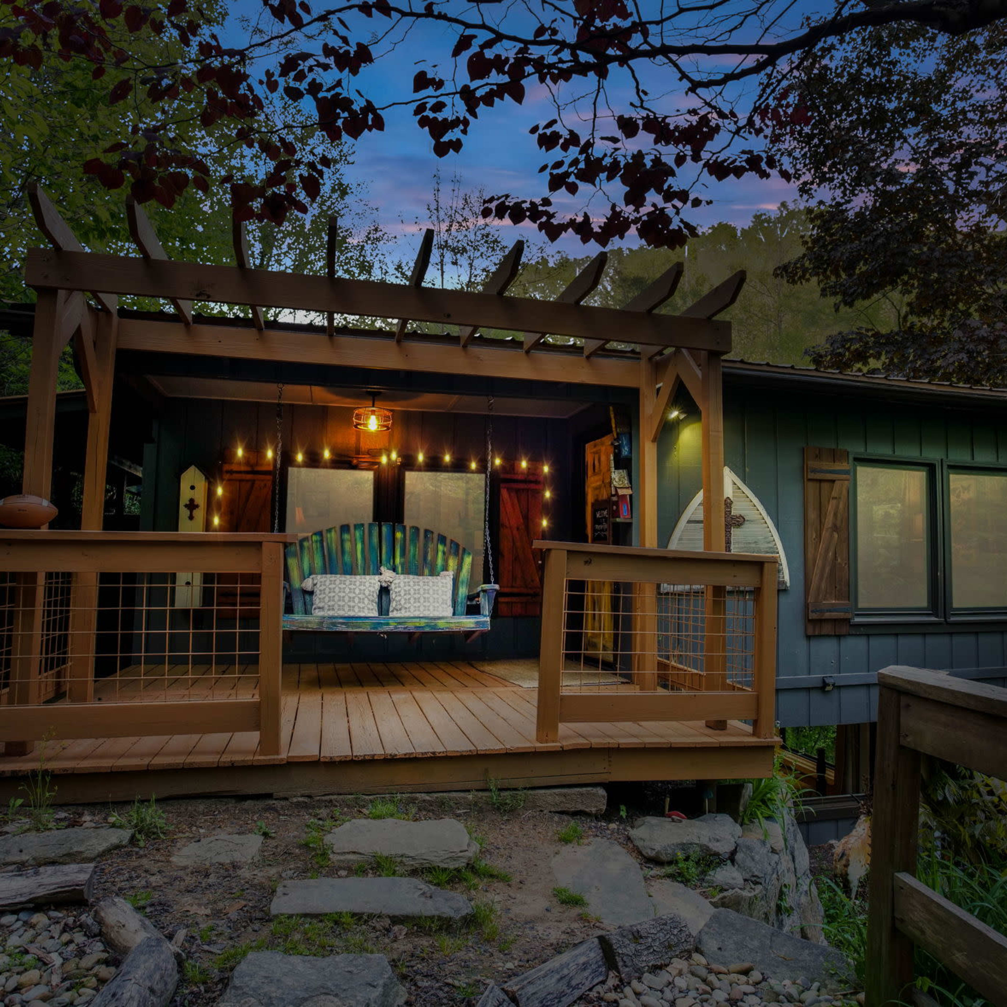 A wooden deck with a covered seating area is attached to a house surrounded by trees and rocks.