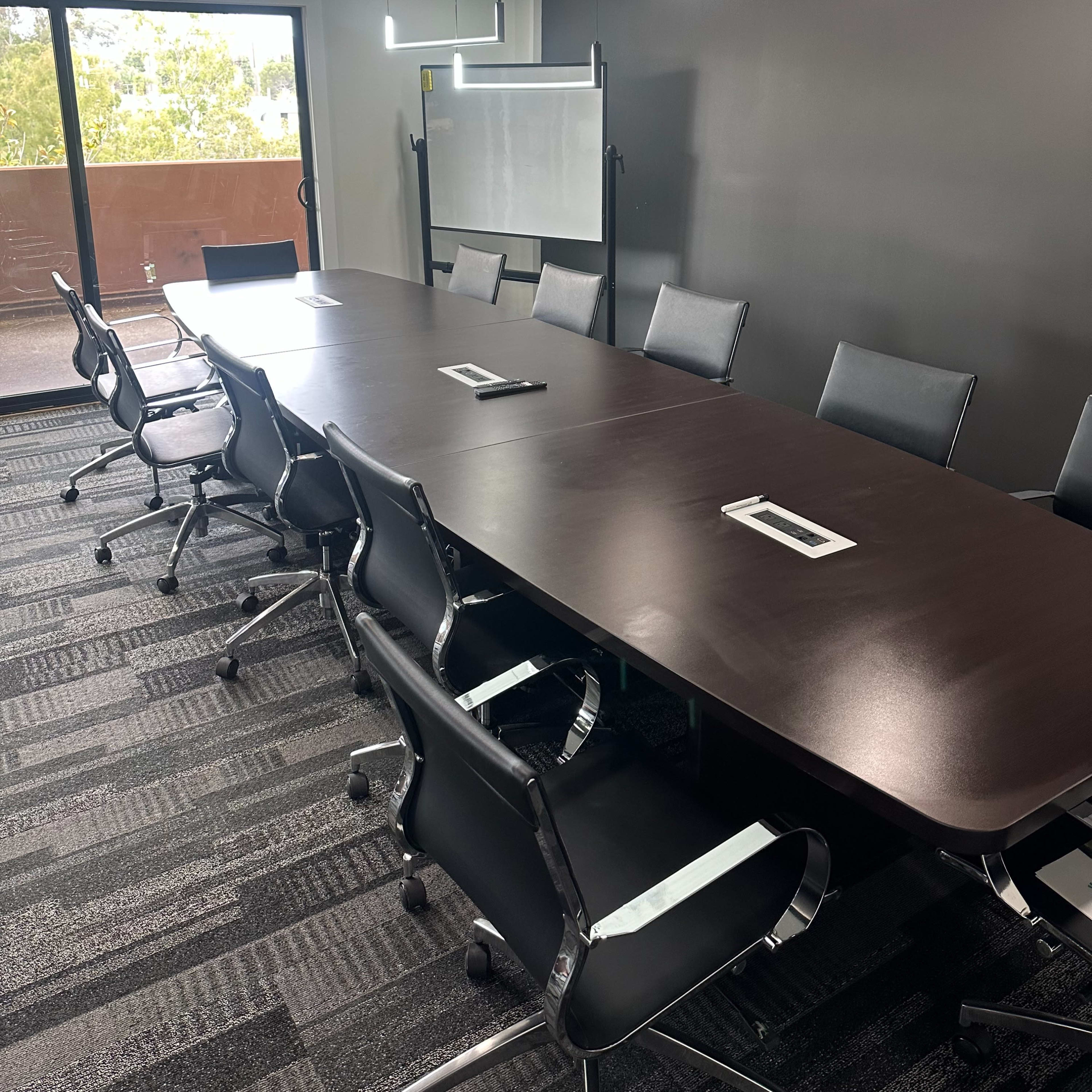 A large conference room features a long wooden table surrounded by black chairs, with a whiteboard on a stand and windows offering natural light.