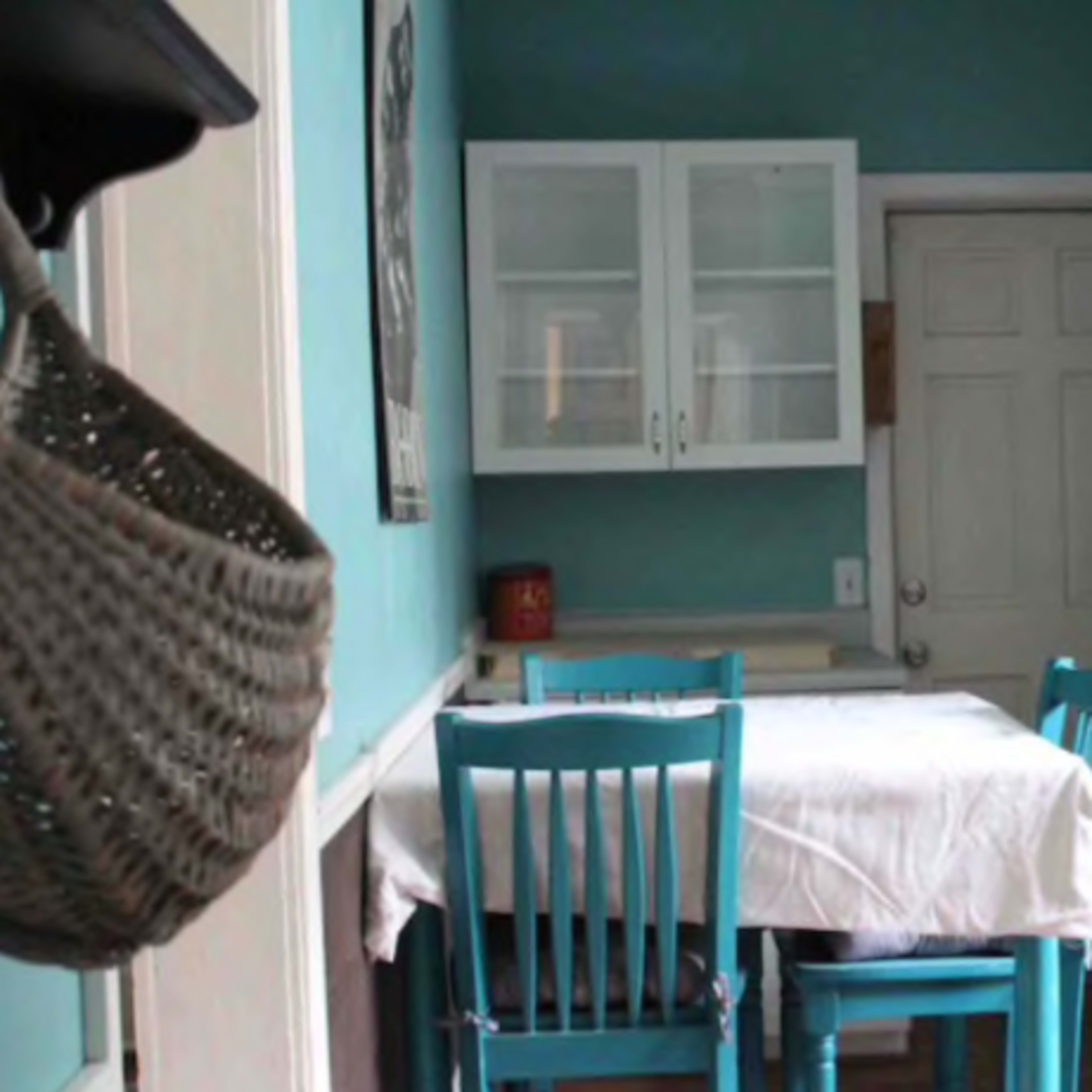 A small kitchen features a table with a white cloth and two blue chairs, with cabinets and a door leading to another room in the background.