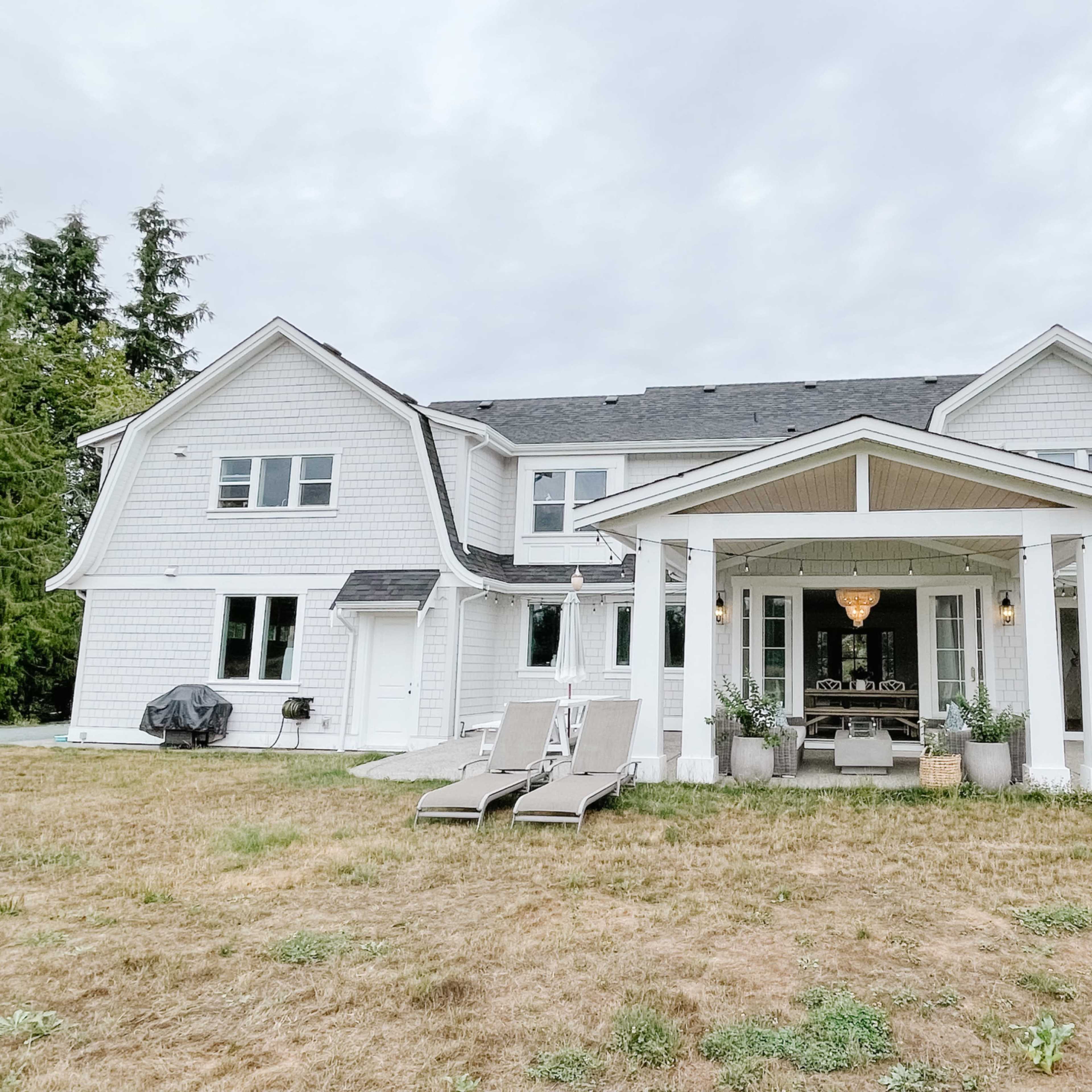 A large white house with a covered porch, featuring outdoor seating and a barbecue grill, sits on a lawn with sparse grass under a cloudy sky.