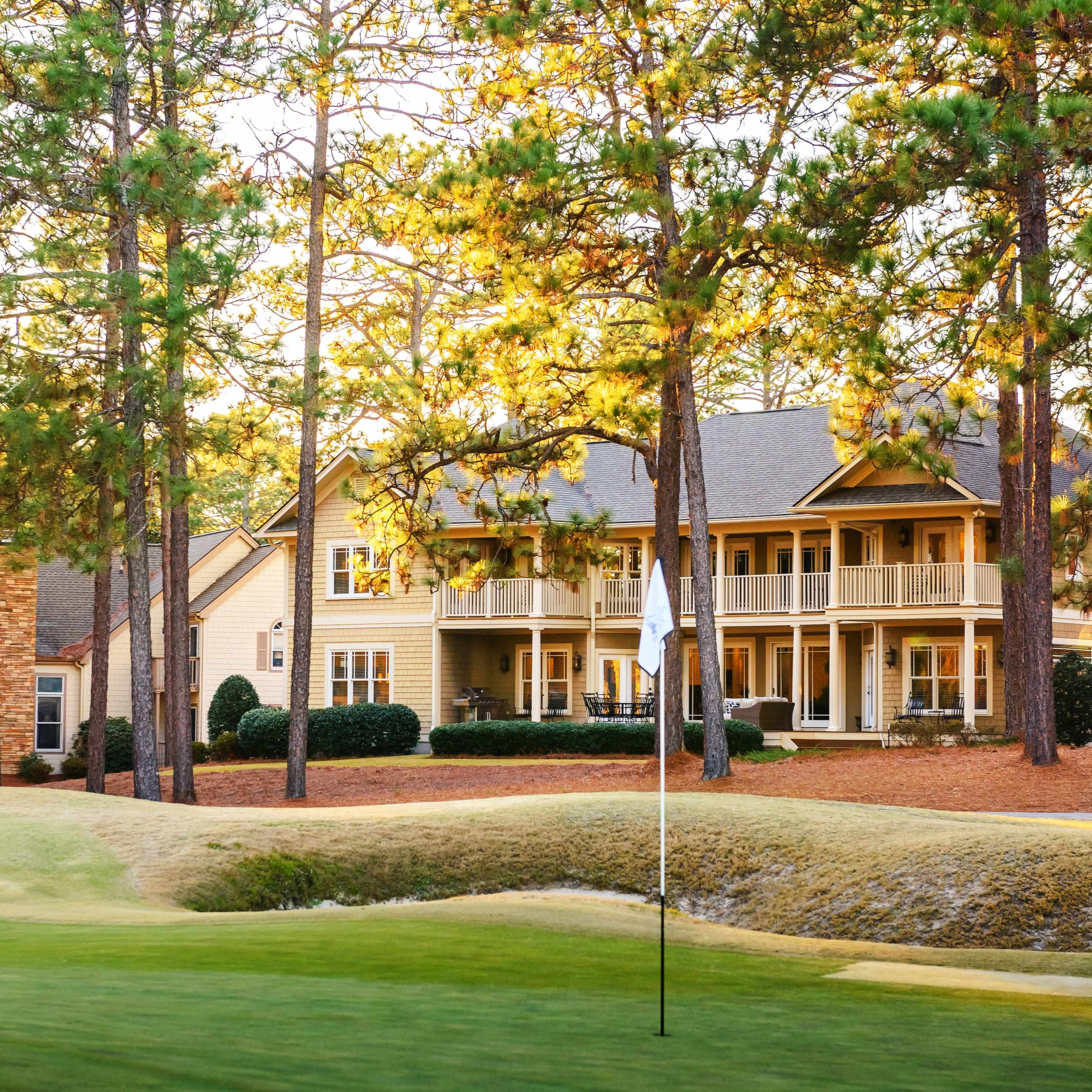 A golf course with a putting green in the foreground and several wooden houses nestled among tall pine trees in the background.