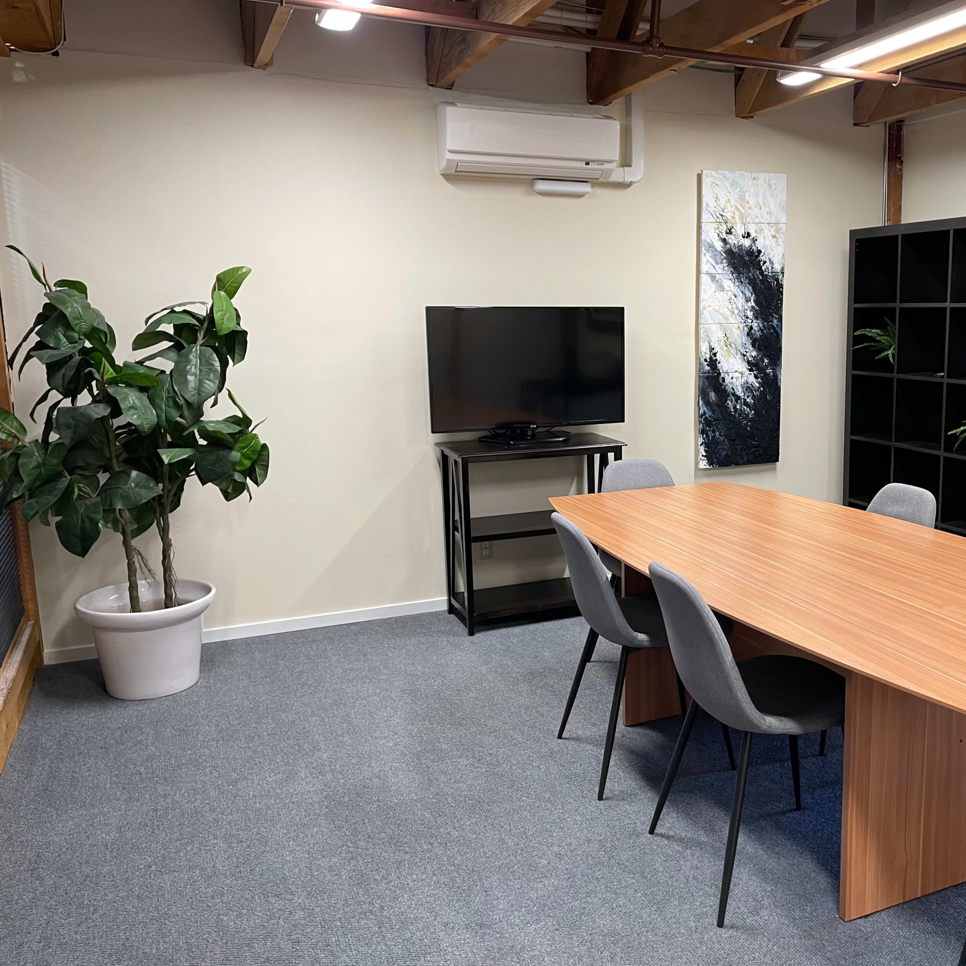 A modern meeting room featuring a wooden table surrounded by six chairs, with a television mounted on the wall and a tall plant in a pot nearby.