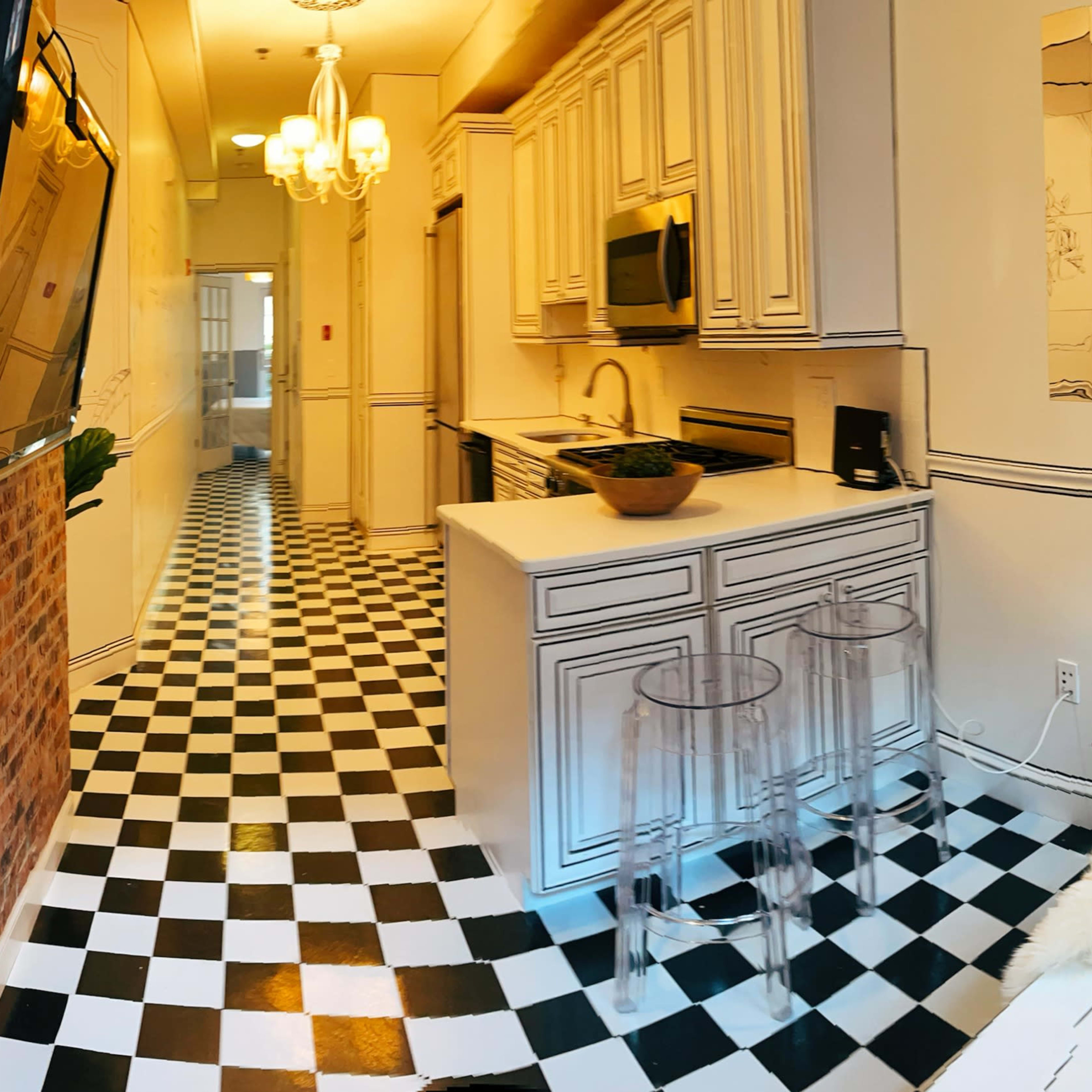 A modern living space with a yellow sofa, checkered black and white flooring, and a kitchen area featuring white cabinetry and a large television mounted on the wall.