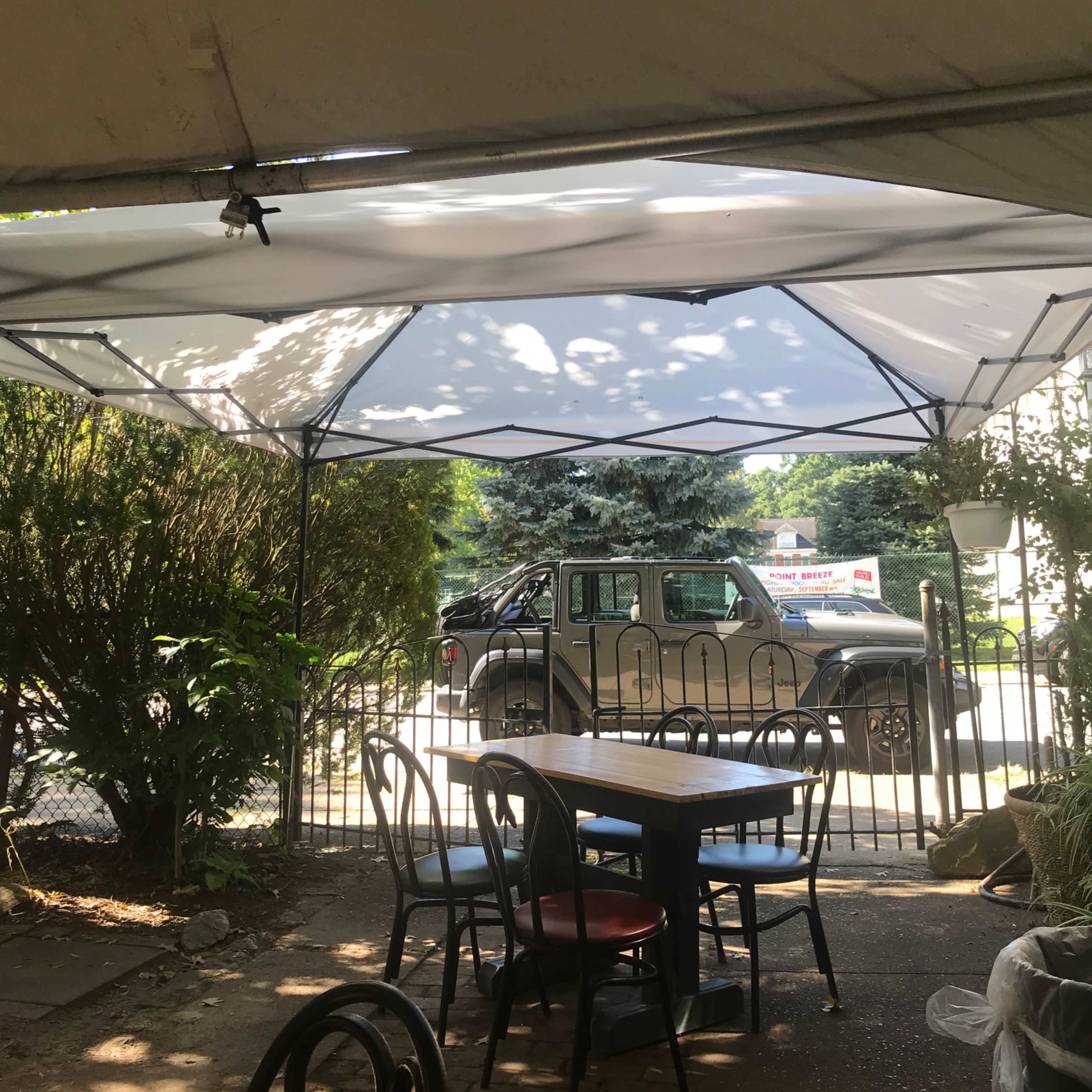 A white canopy is set up over a table and chairs in a shaded outdoor area, with a parked vehicle visible through an open gate in the background.