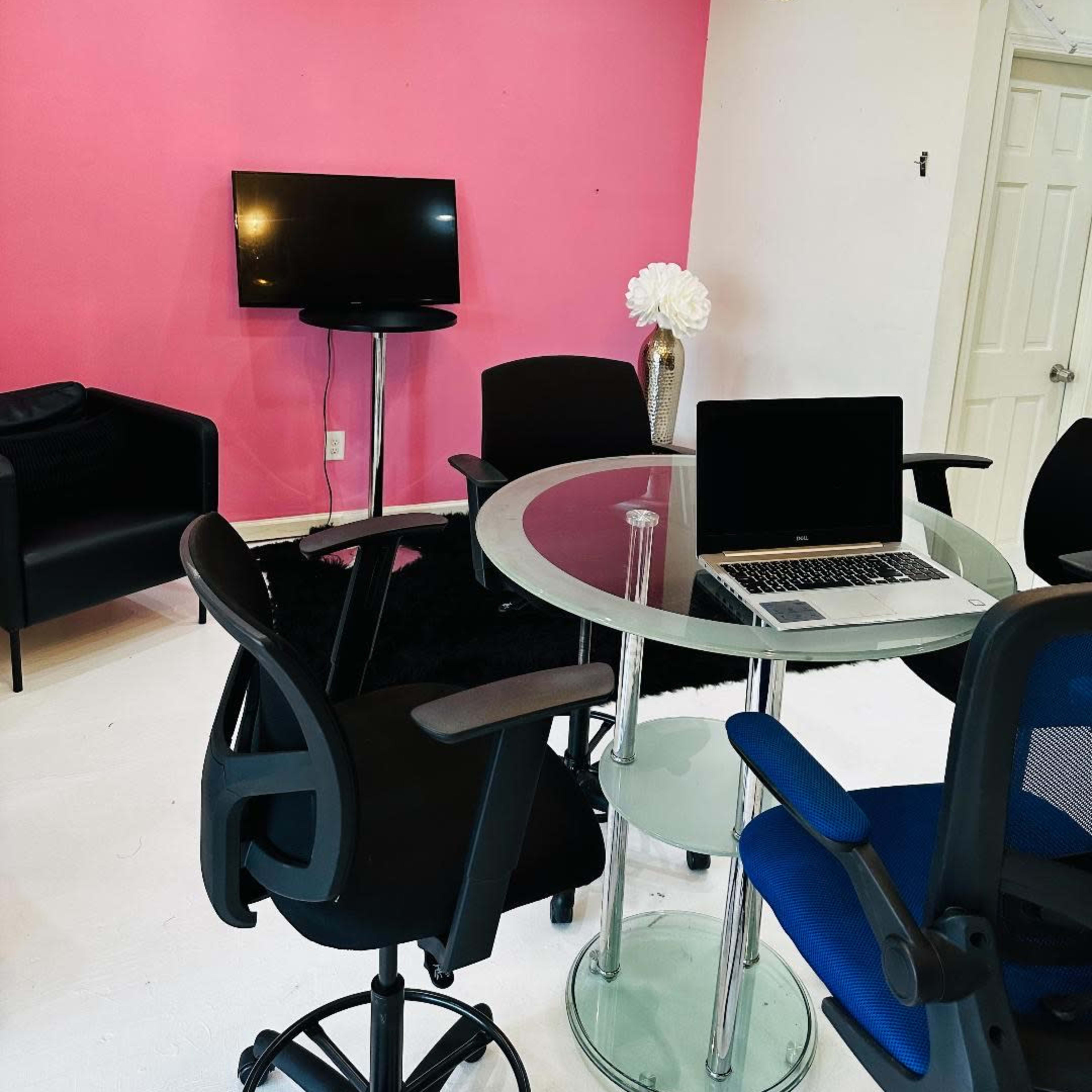 A modern office space with a glass table, several chairs, a television mounted on a pink wall, and a chandelier overhead.