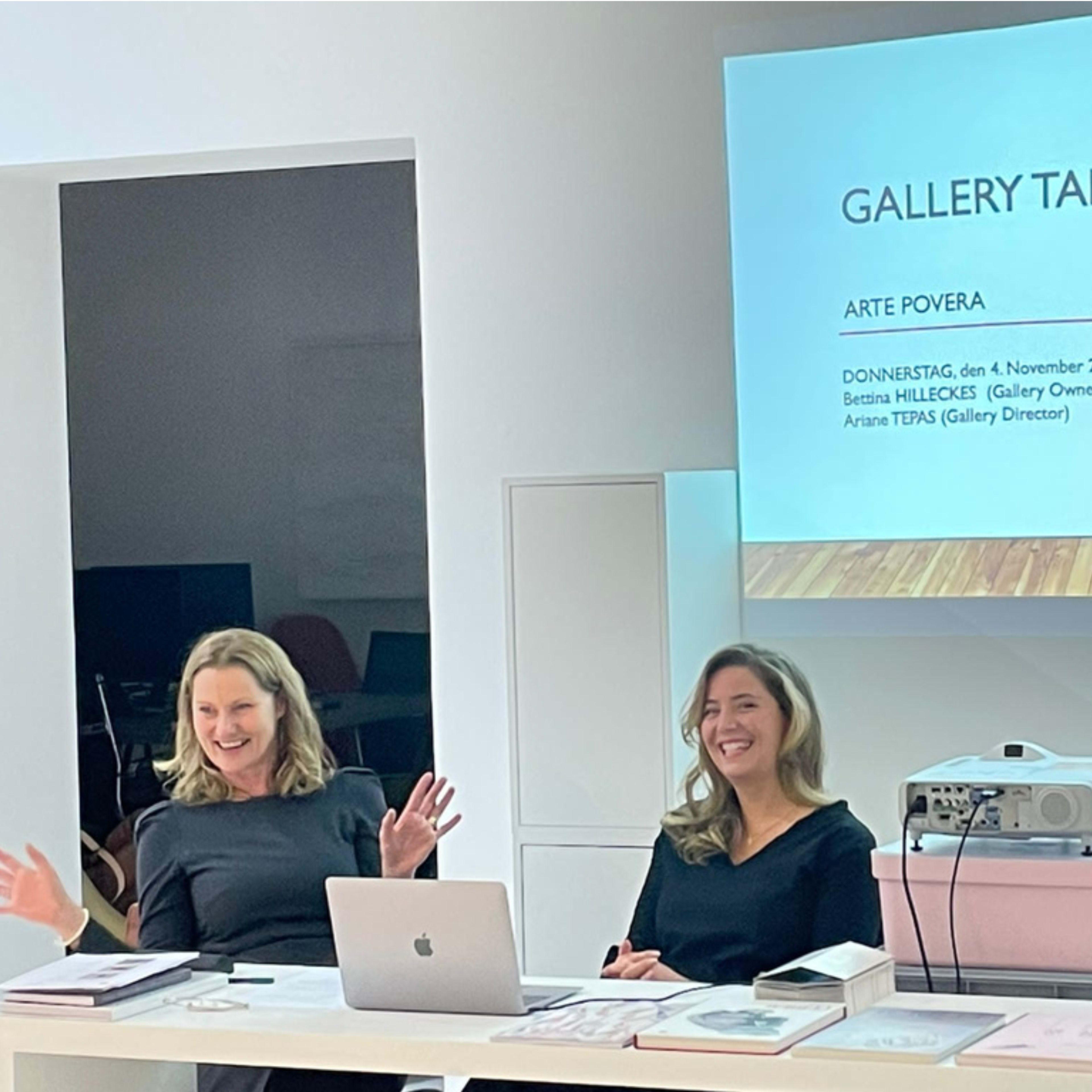 Two women are seated at a table during a gallery talk event with a presentation slide about "Arte Povera" displayed behind them.
