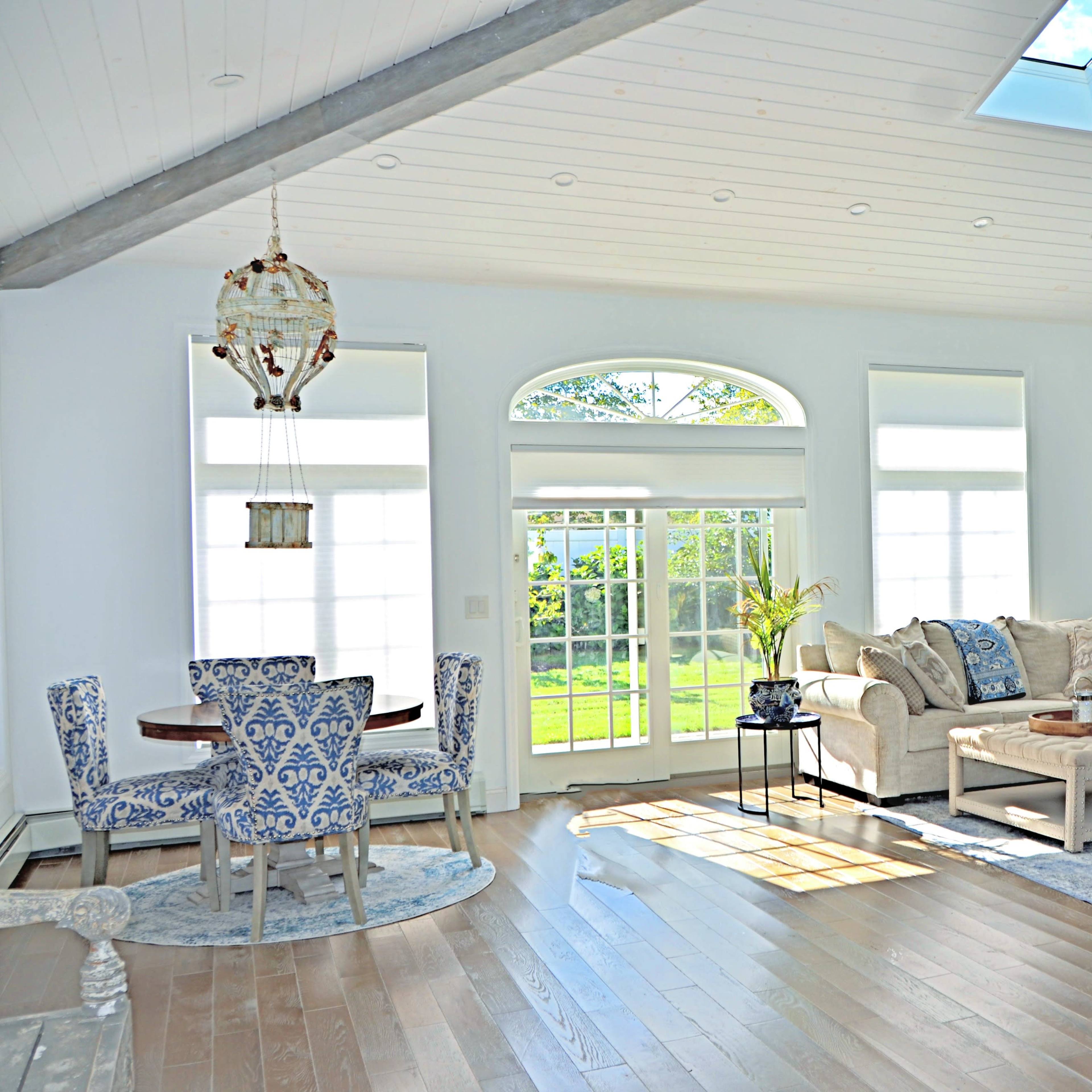 A bright living room features a comfortable sofa, a wooden coffee table, and a dining set, illuminated by natural light streaming through large windows.