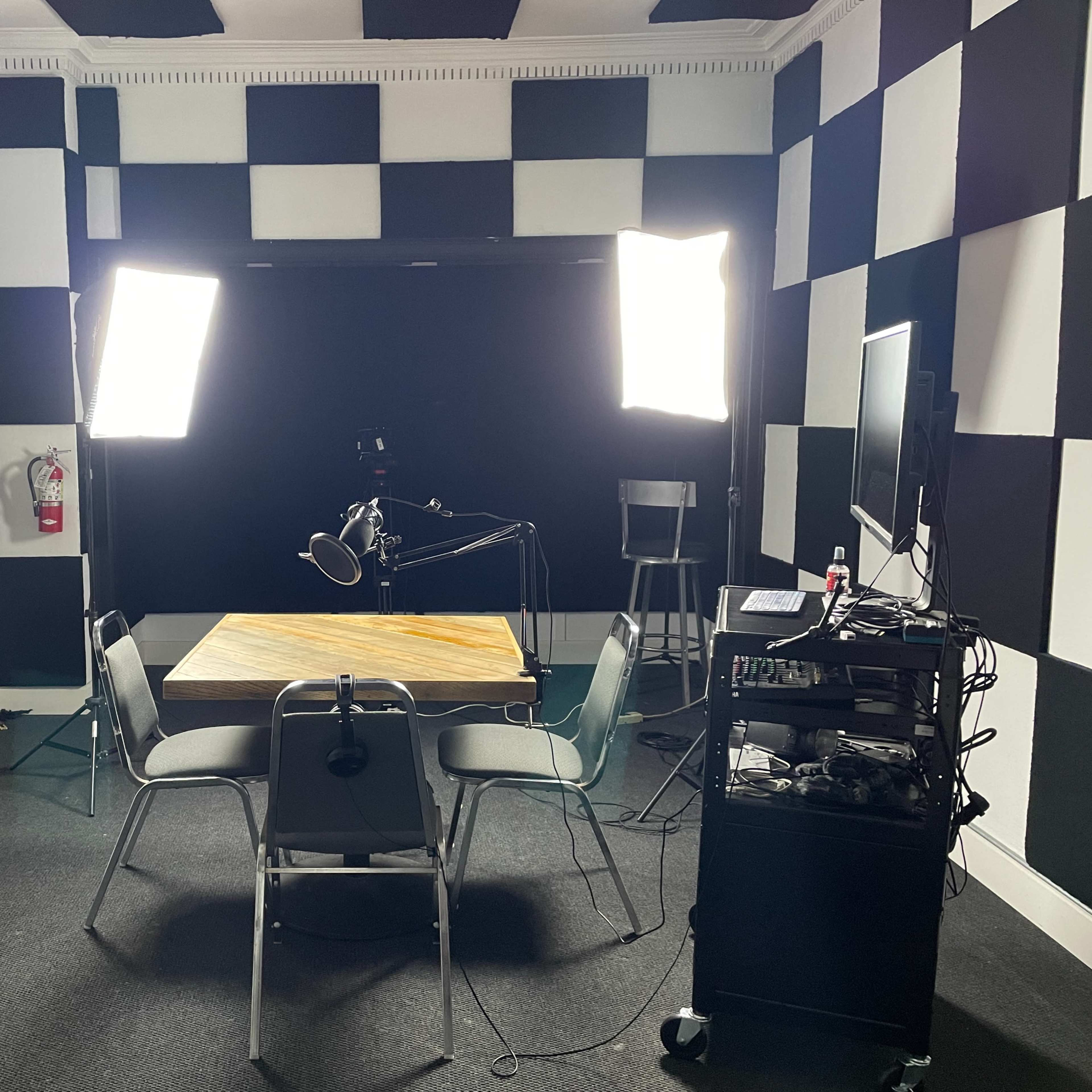 A recording studio features a central table surrounded by chairs, with two bright lights positioned to illuminate a black backdrop and various equipment on a nearby cart.