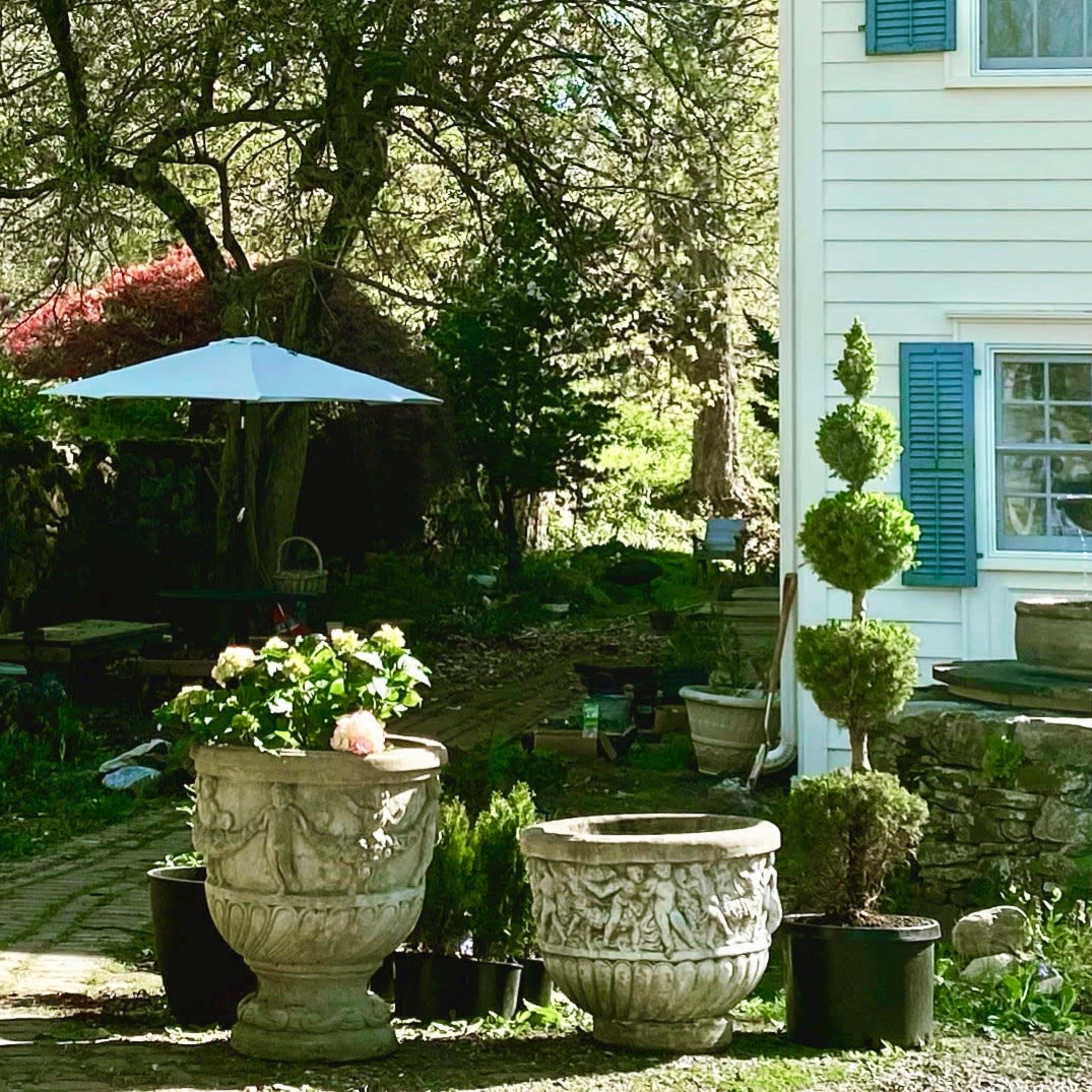 A tiered stone fountain stands next to two decorative planters in a garden area beside a white house.
