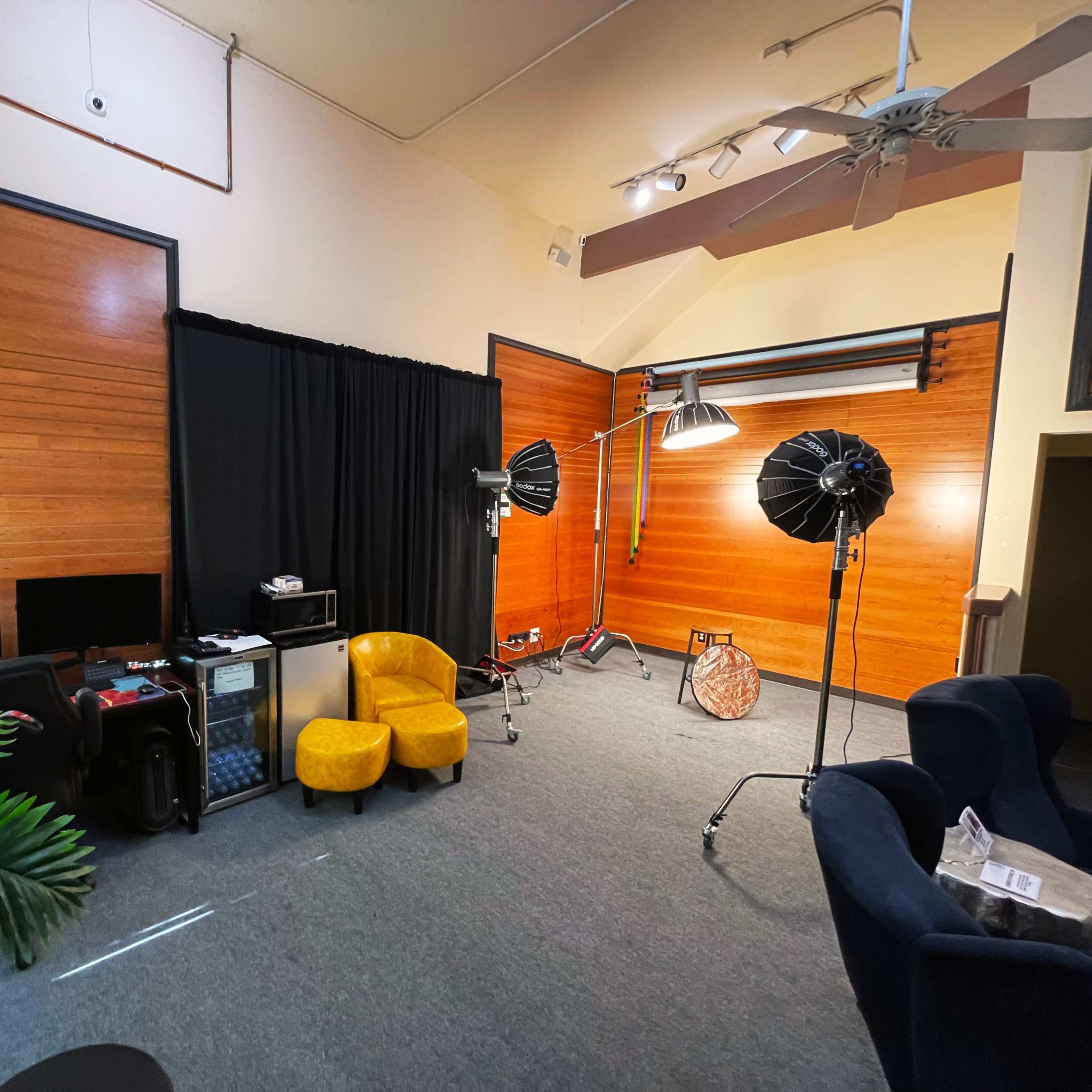 A photography studio featuring wooden walls, a black backdrop, two light stands with softboxes, and a pair of yellow armchairs.