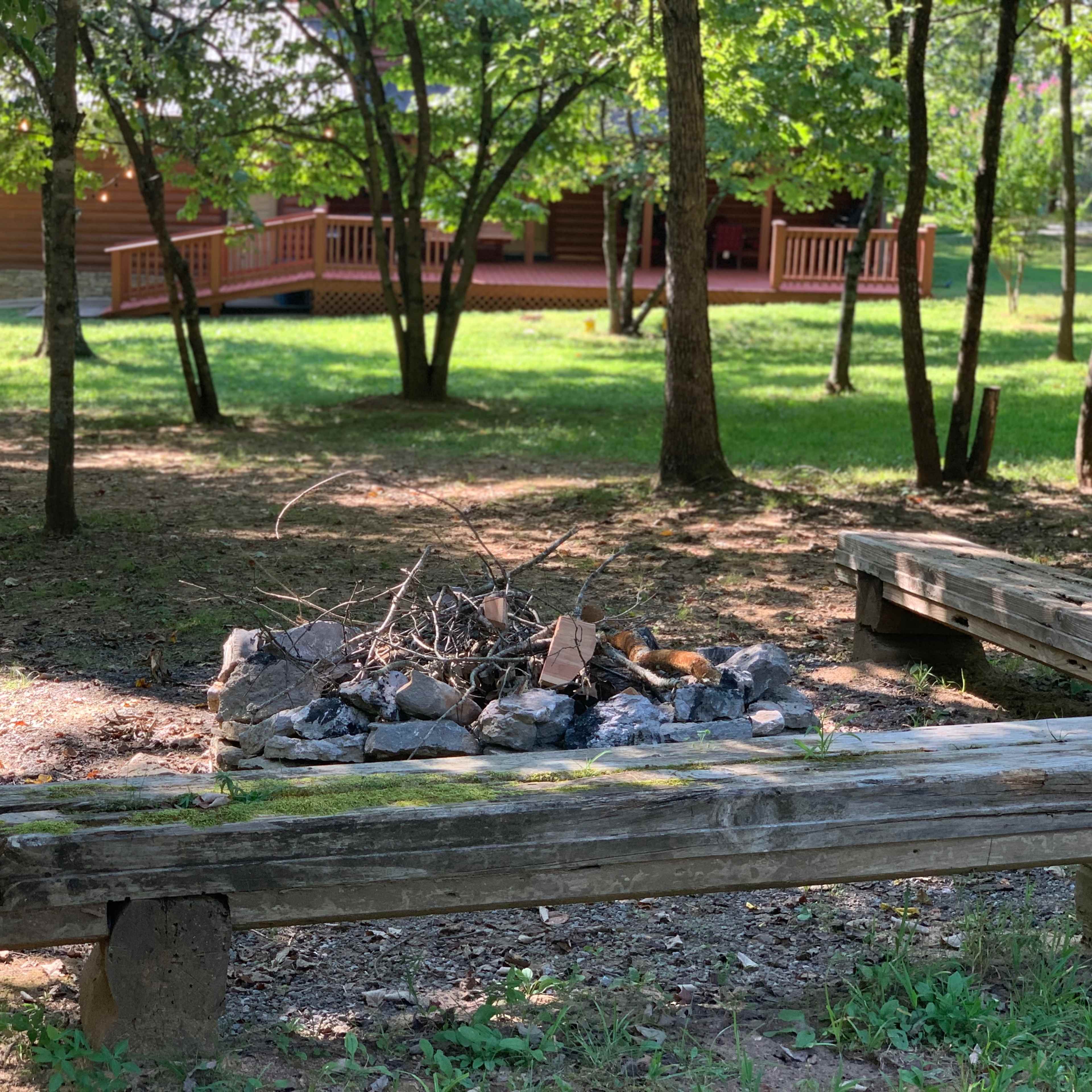 A circular fire pit made of stones surrounded by wooden benches is located in a grassy area with trees and a cabin in the background.