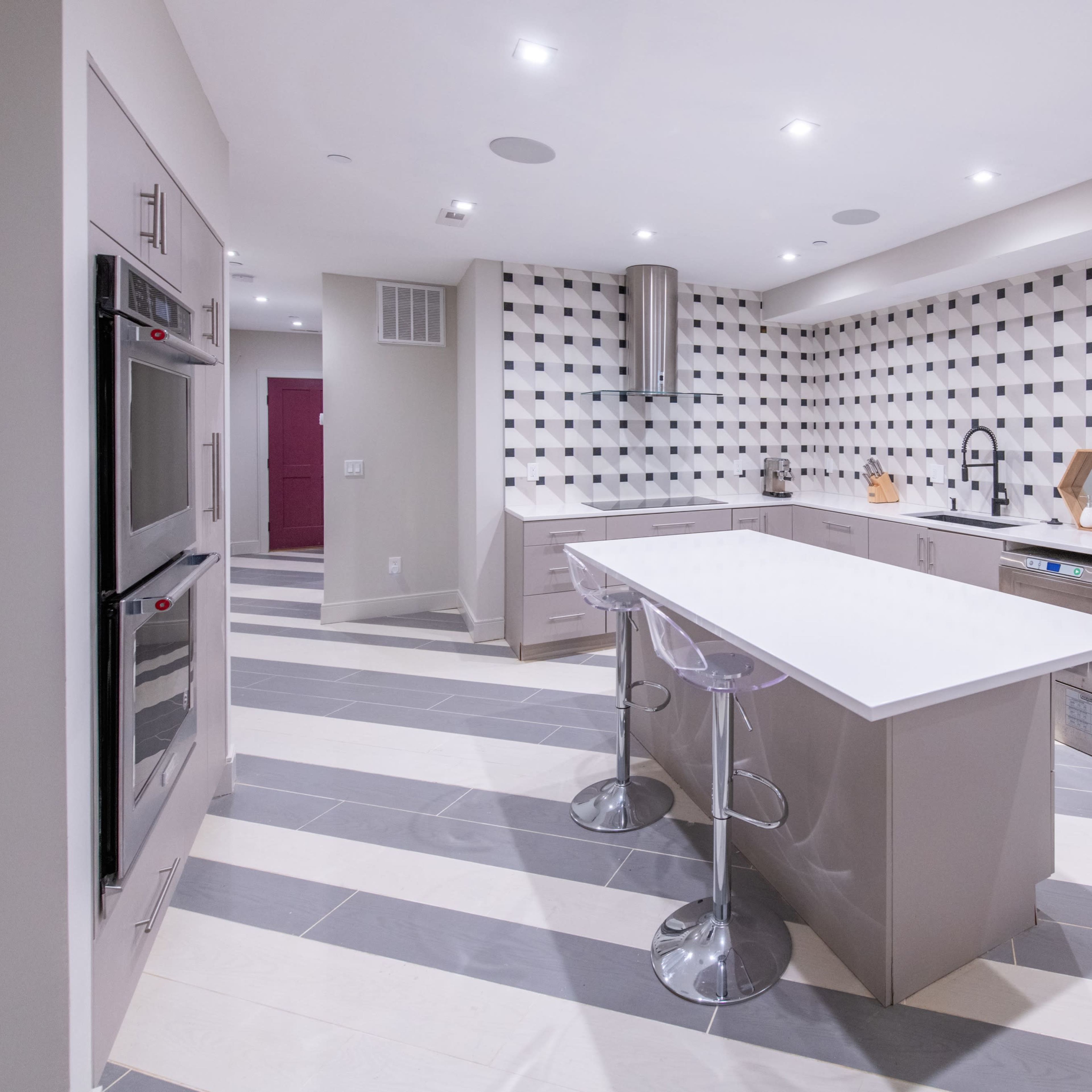 A modern kitchen featuring a central island with two clear stools, stainless steel appliances, and a geometric patterned backsplash.
