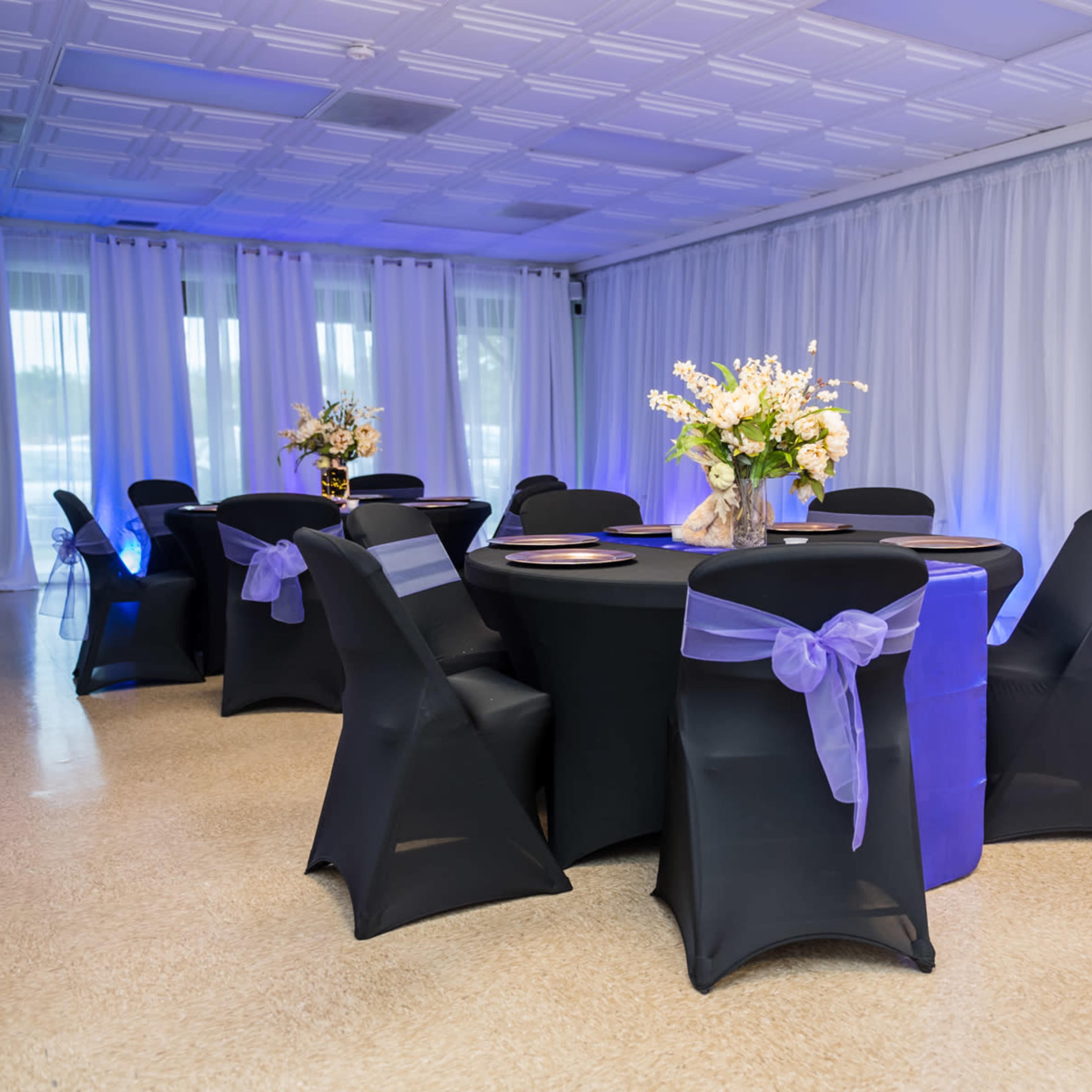 A banquet room features round tables with black tablecloths and lavender sashes, adorned with floral centerpieces, against a backdrop of white drapes.