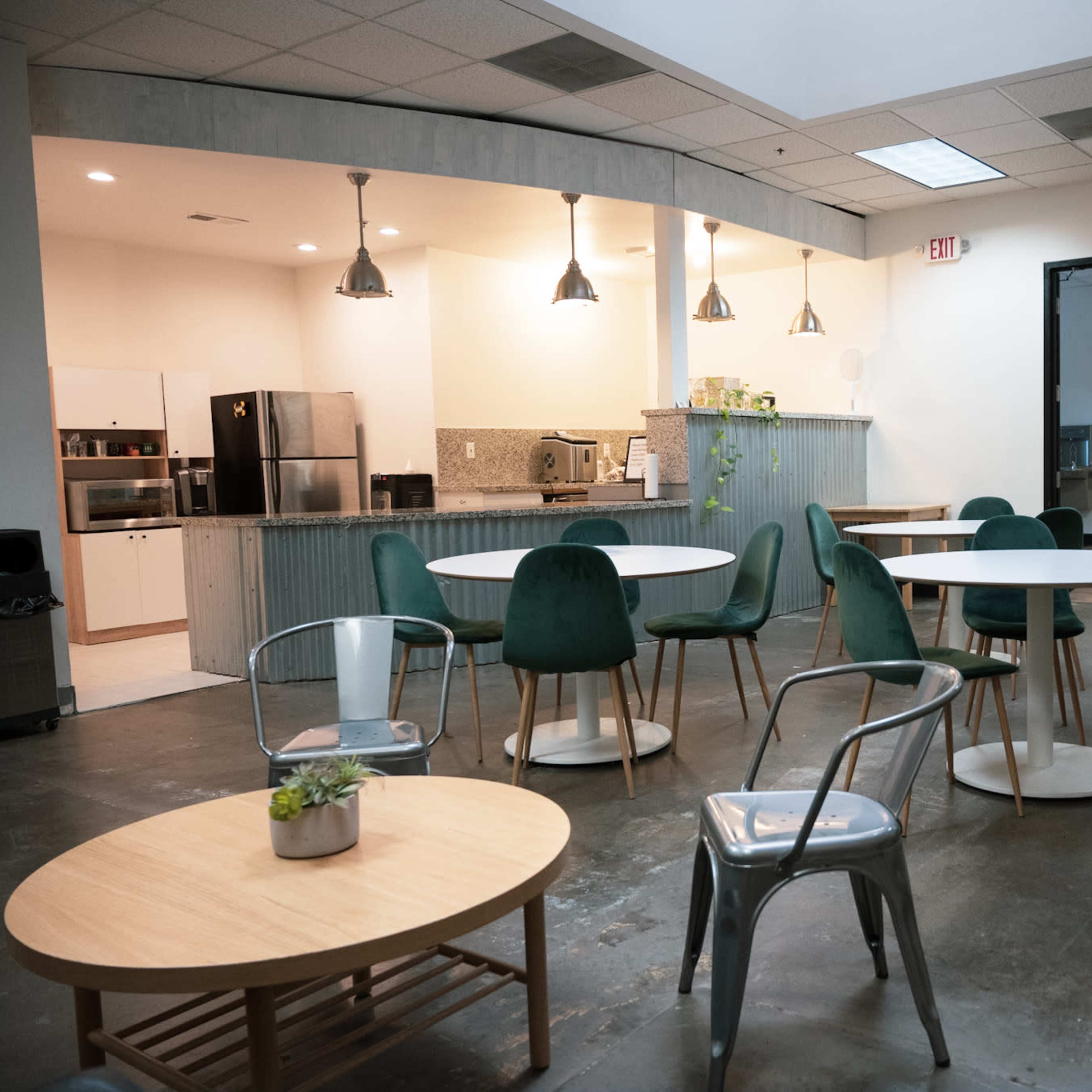 A modern break room with several tables and chairs, a kitchenette in the background, and various kitchen appliances visible.