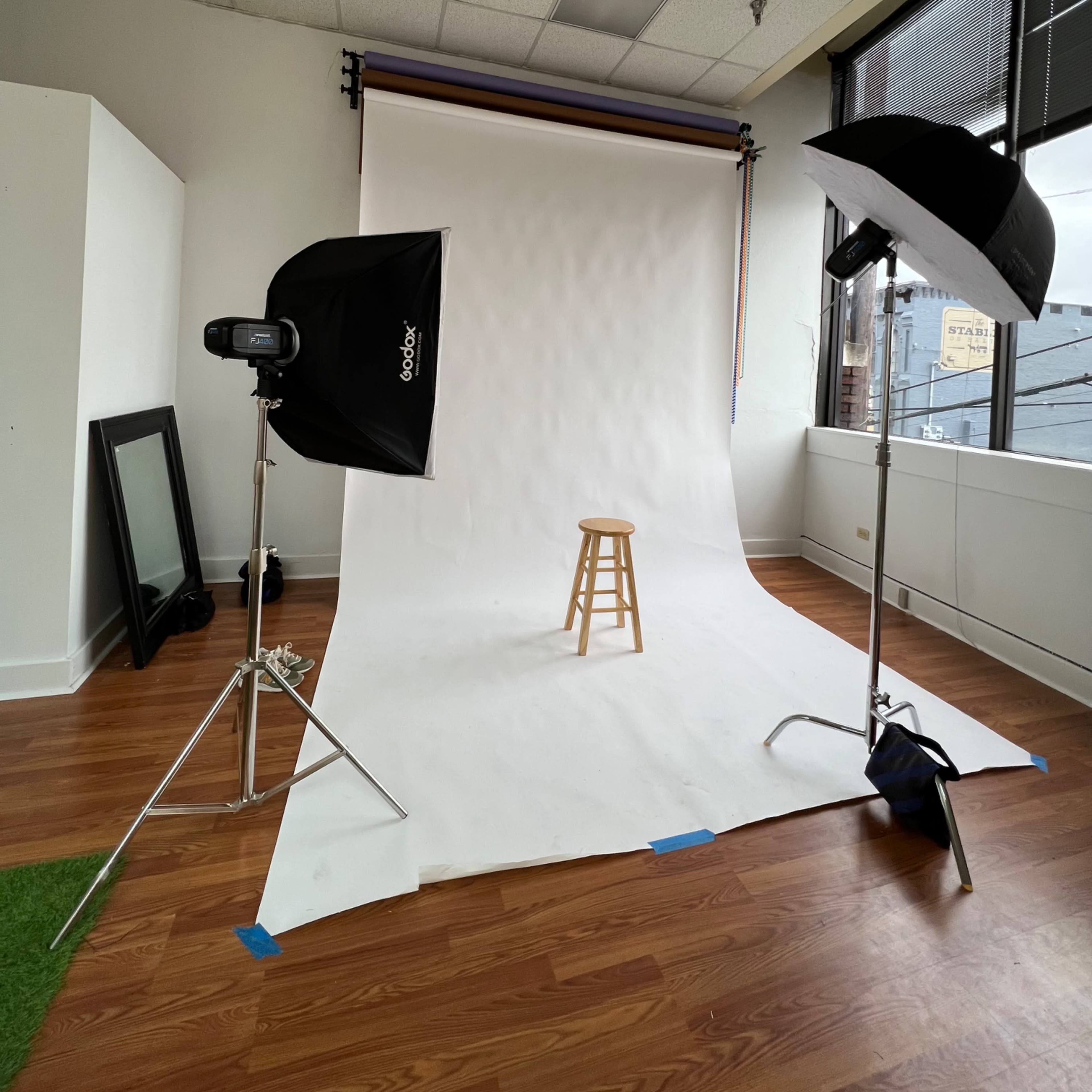 A photography studio with a white backdrop, a wooden stool in the center, and two light sources positioned on either side.