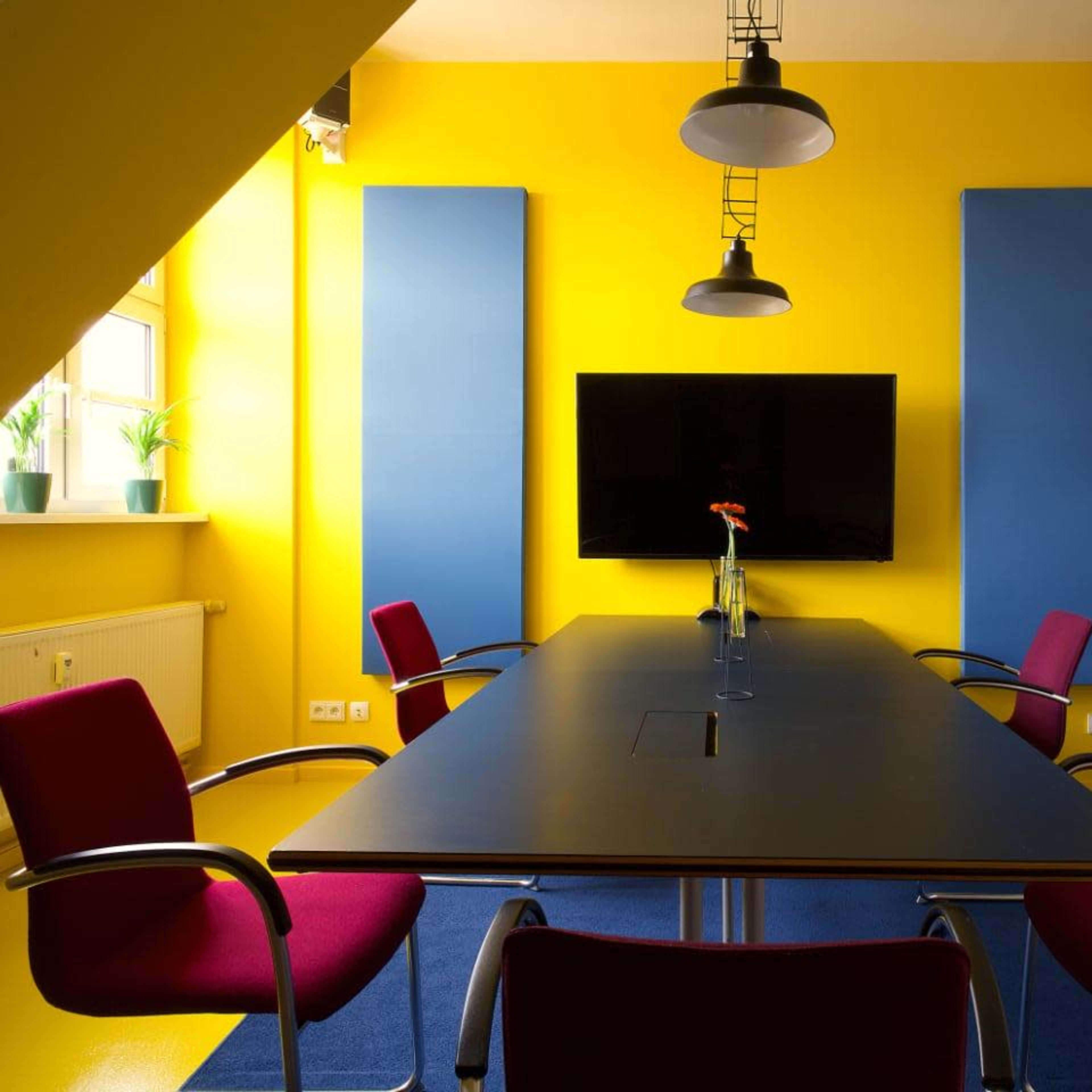 A modern conference room features a black table surrounded by red chairs, a large screen on the wall, and bright yellow walls with sound-absorbing panels.