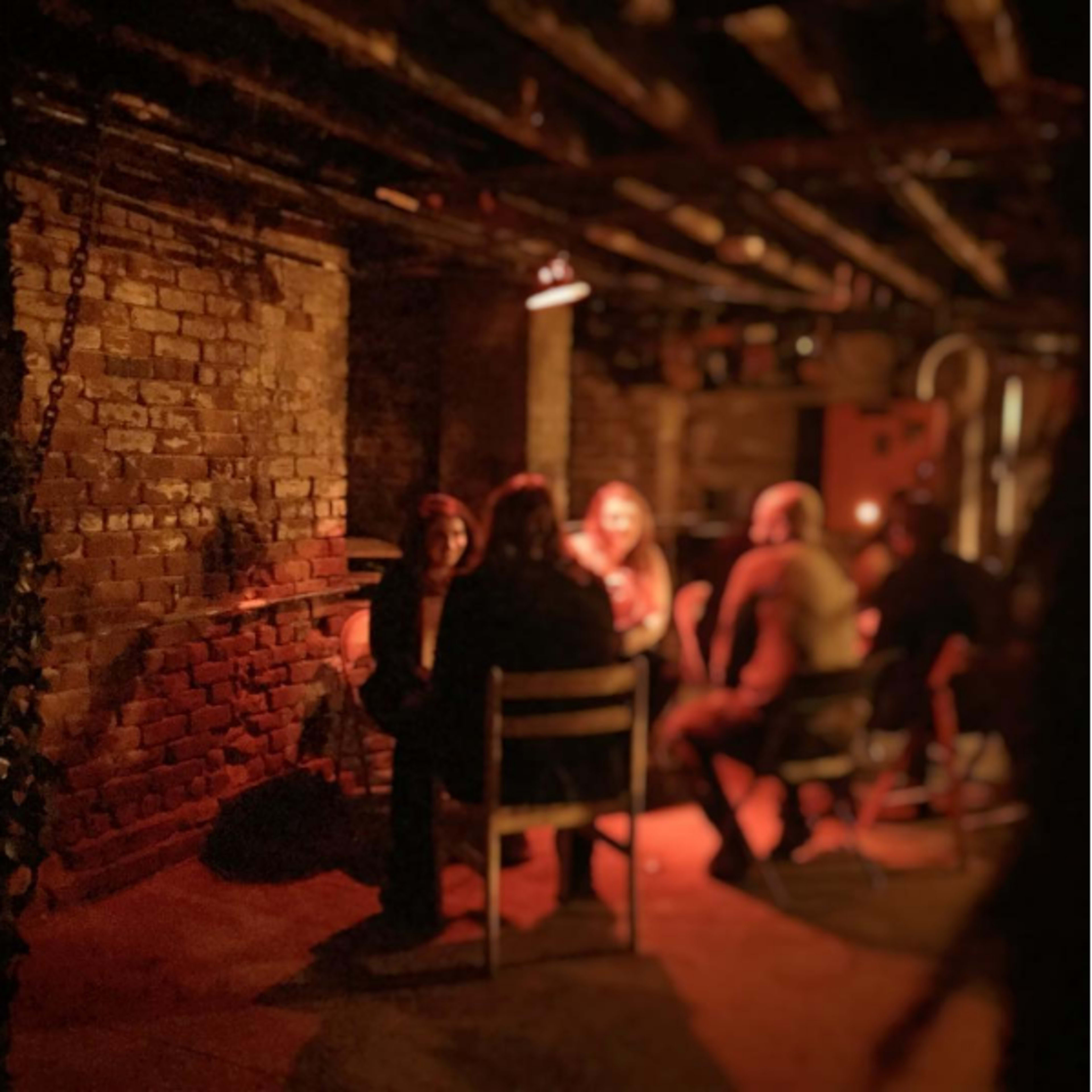 A group of people sits around a table in a dimly lit basement with brick walls.
