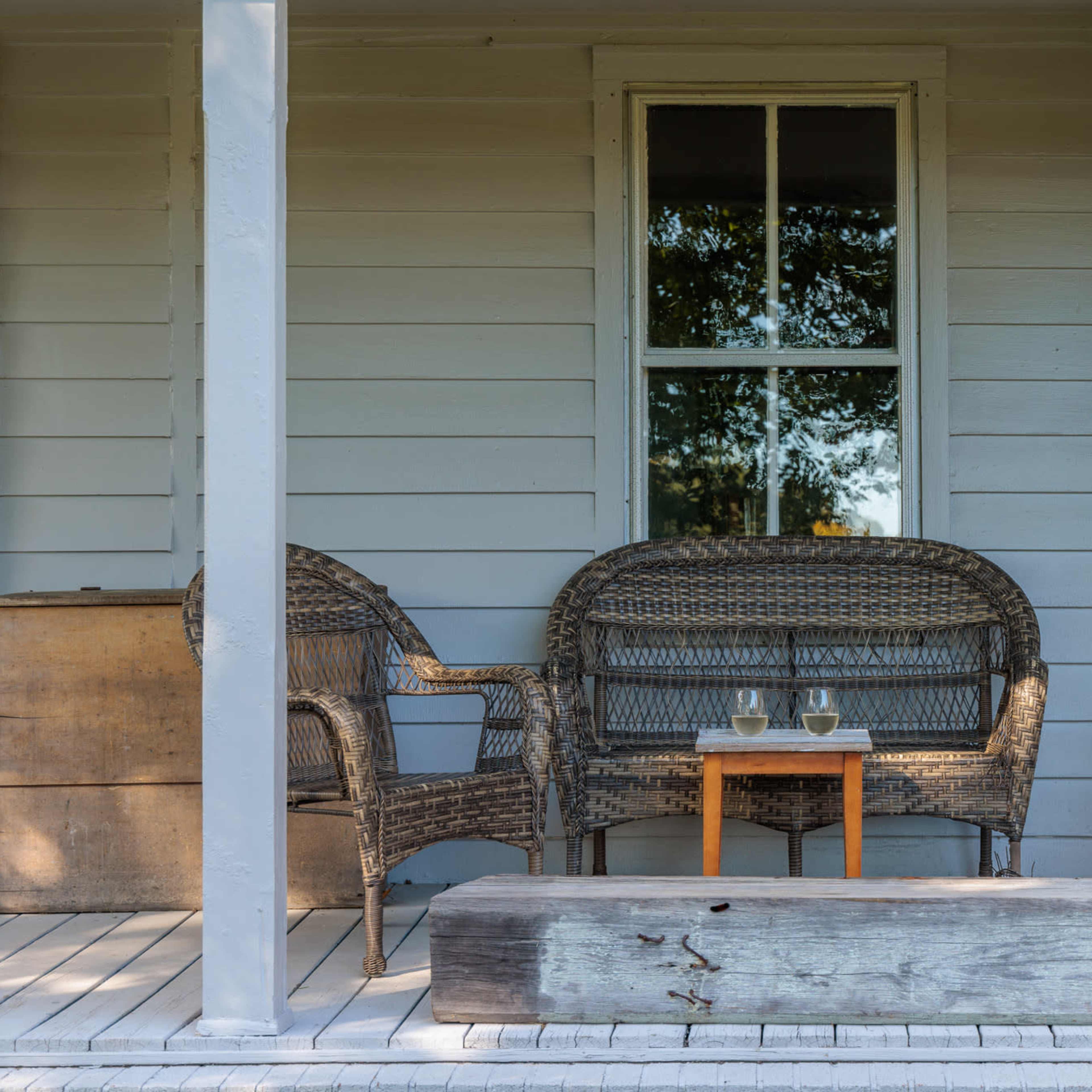 A porch features two woven chairs and a small table with two glasses, situated in front of a house with a large window.