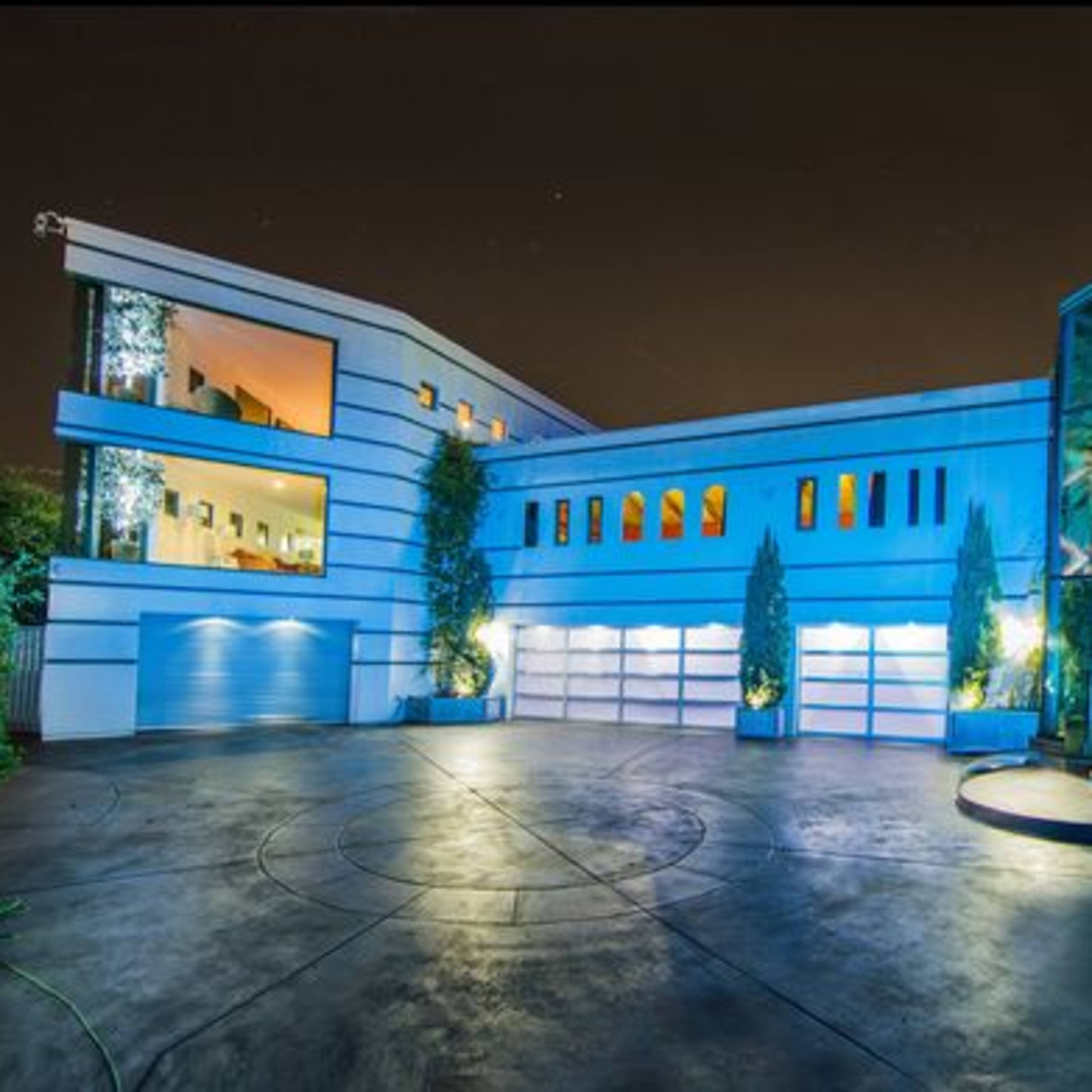 A modern residential building with a glass entrance, surrounded by landscaped gardens and palm trees, illuminated at night.