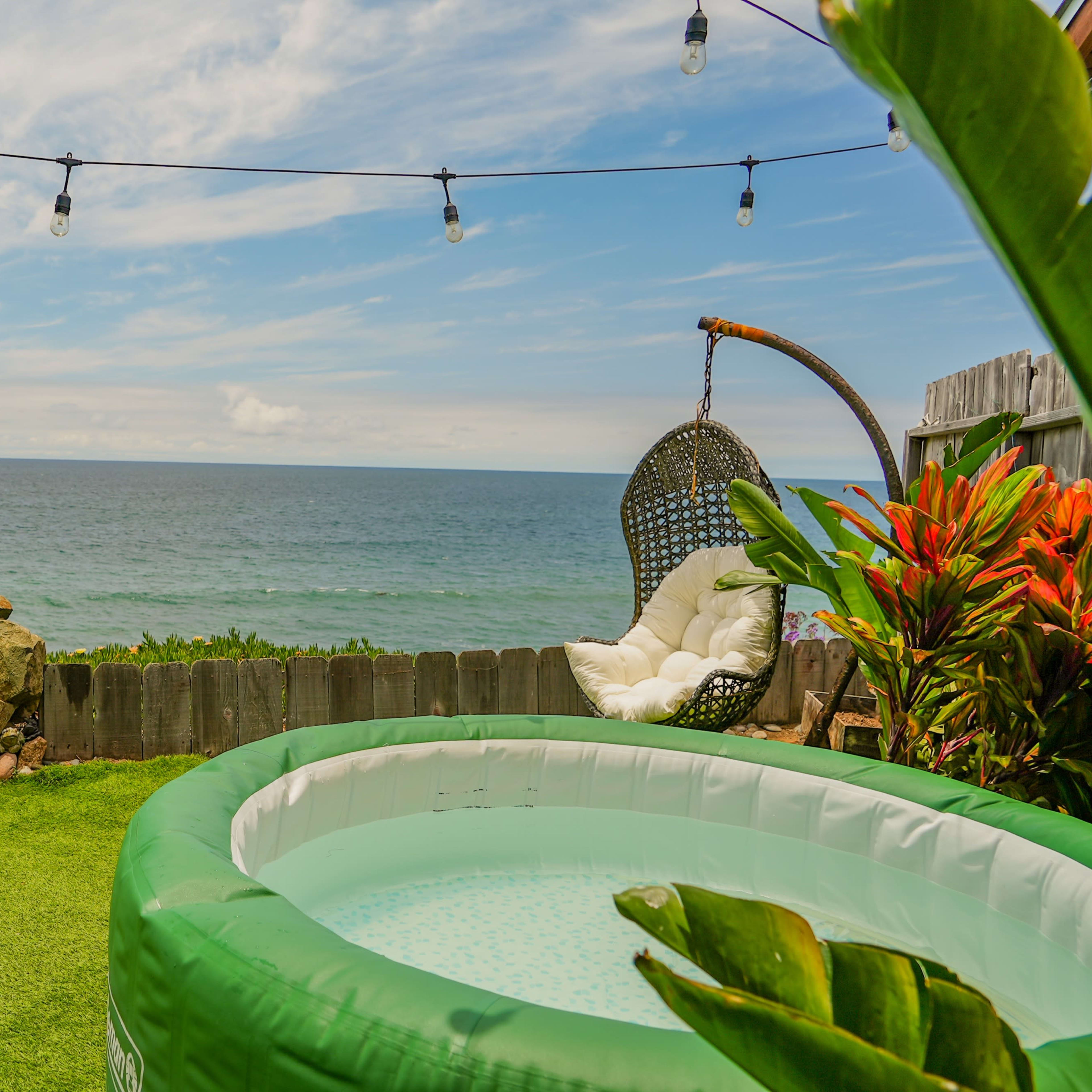 A backyard with a small inflatable pool and a hanging chair, overlooking the ocean under a sky with scattered clouds.